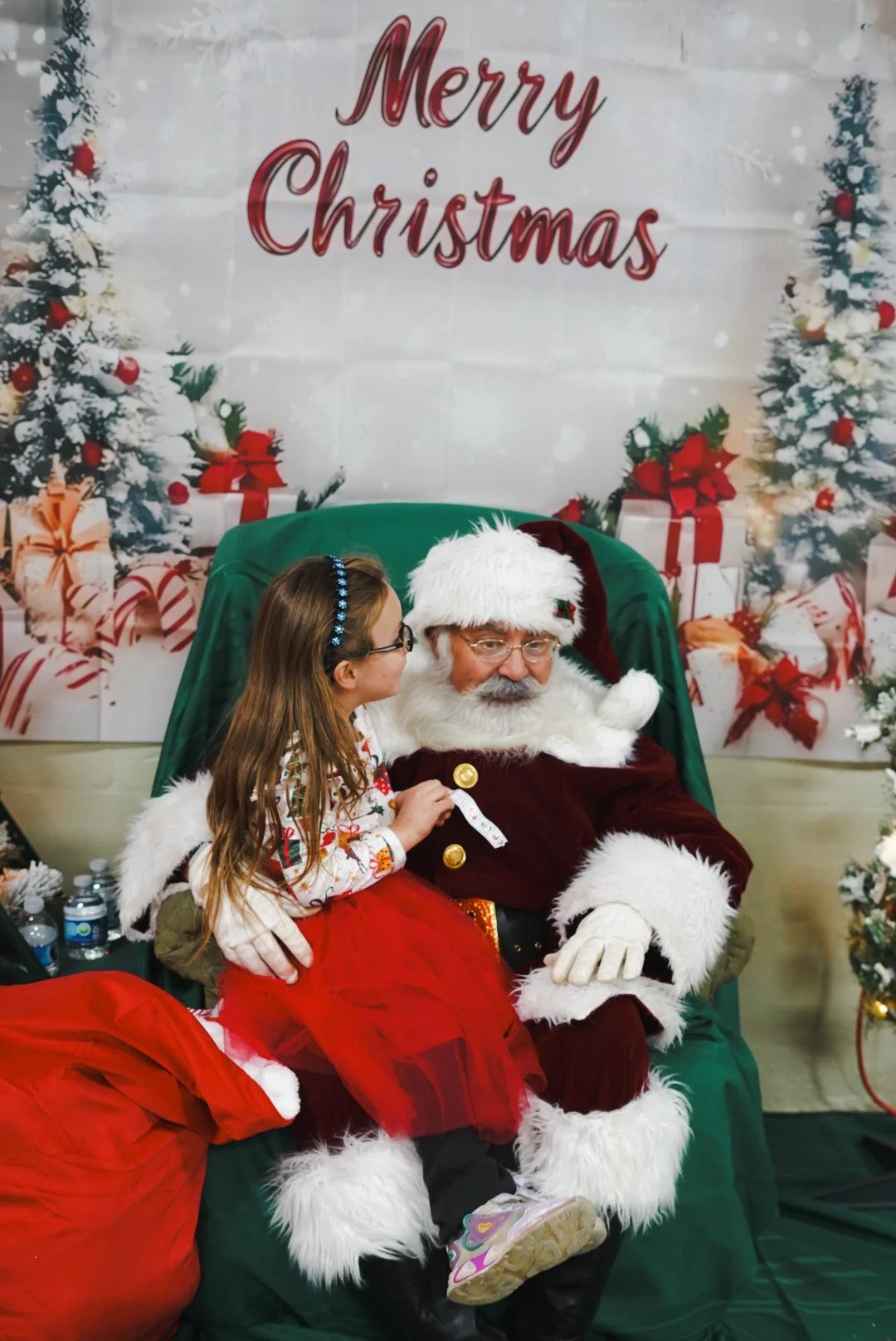 A young girl sits on Santa Claus's lap, talking to him in front of a festive Christmas backdrop with the message "Merry Christmas" and decorated trees.