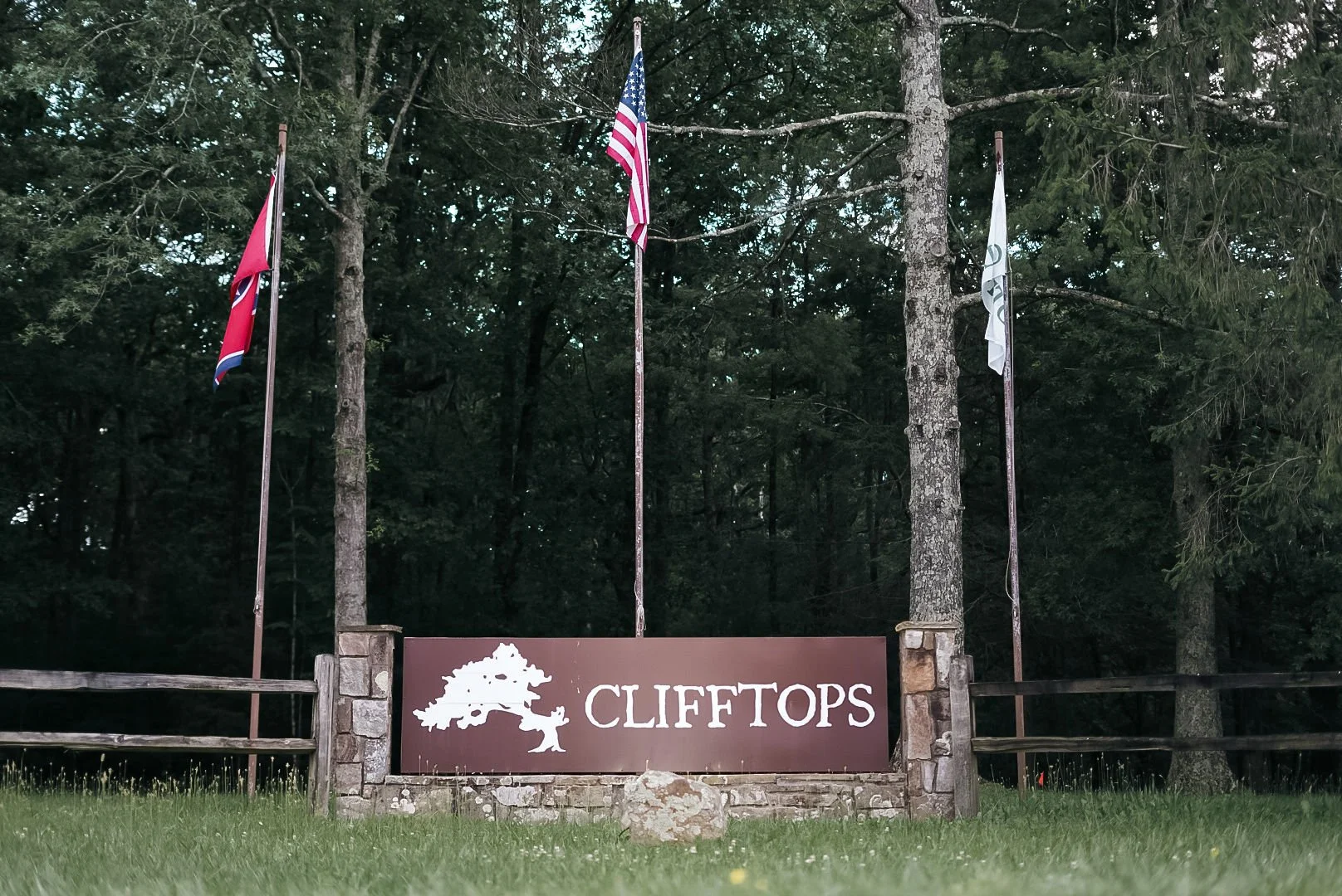 Sign for Clifftops with a tree graphic, surrounded by trees and four flags, including the American flag, in a wooded area.