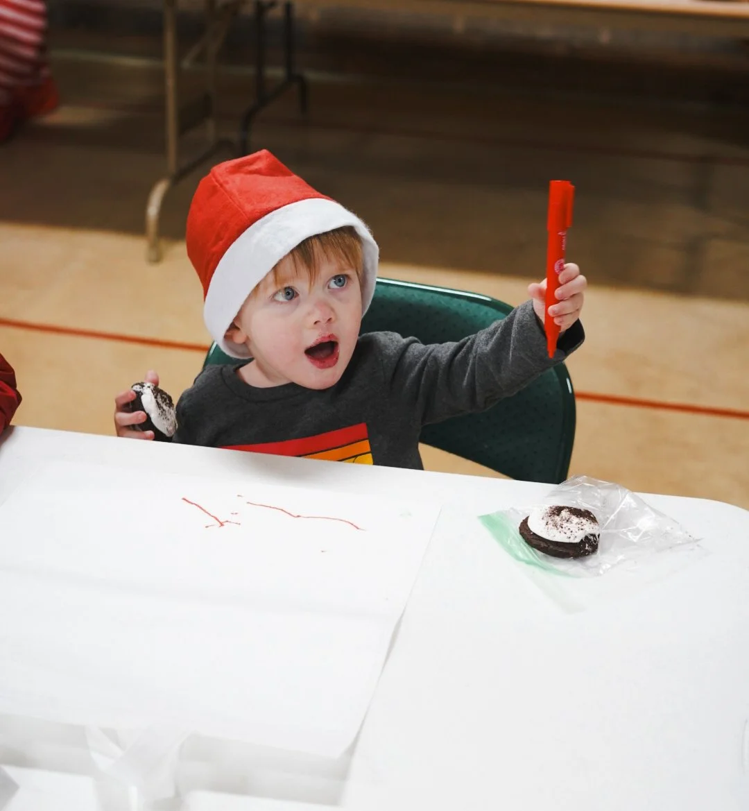 Child wearing a Santa hat sitting at a table with a white tablecloth, holding a red marker, and an Oreo cookie on the table.