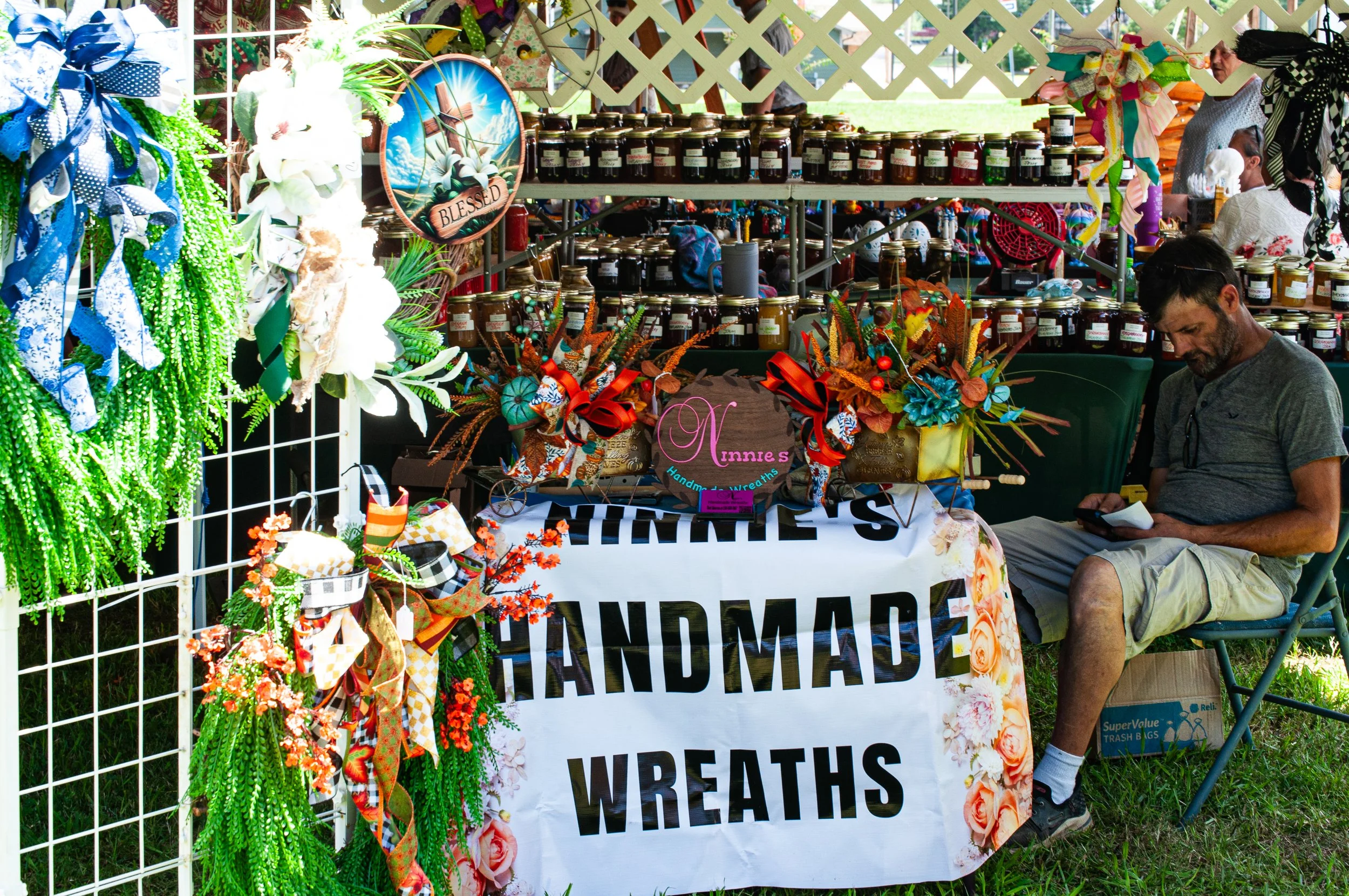 A vendor stand at an outdoor market selling handmade wreaths and wreath accessories, decorated with colorful ribbons and flowers. A man is sitting on a chair looking at his phone next to the stand.
