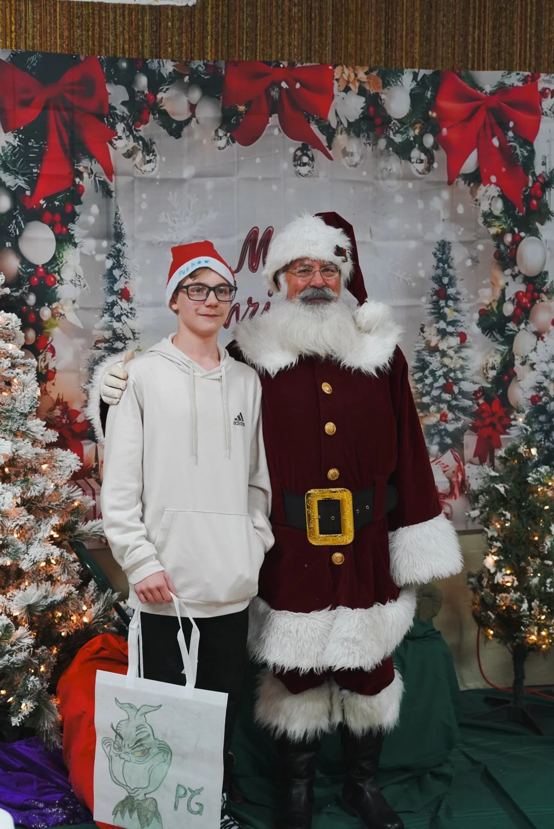 A young person with glasses and a Santa hat posing next to Santa Claus, who is dressed in a traditional red and white suit, in front of a Christmas-themed background with decorated Christmas trees and festive decorations.