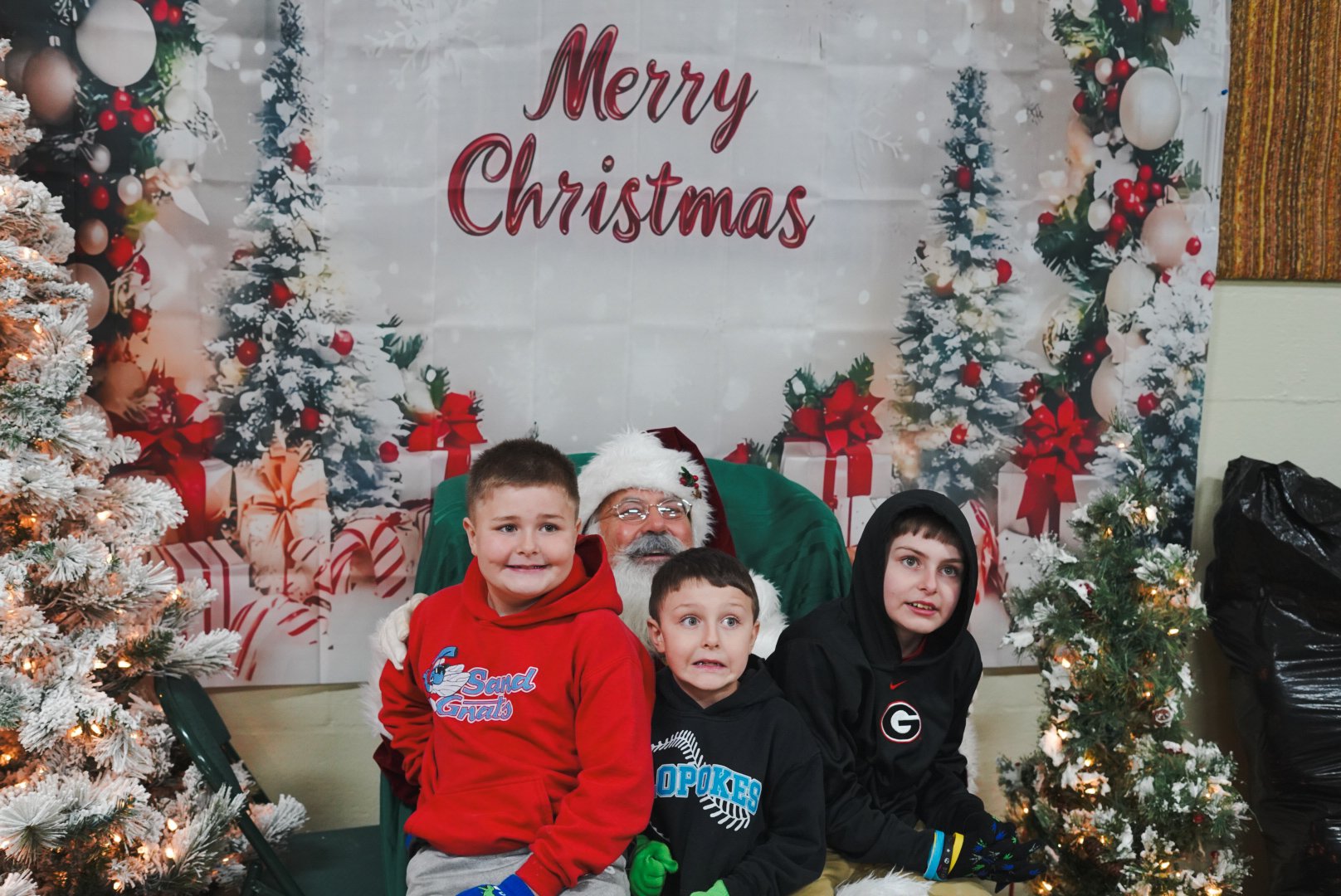 A group of three children and a man dressed as Santa Claus posing for a photo during Christmas, with decorated Christmas trees and a 'Merry Christmas' banner in the background.