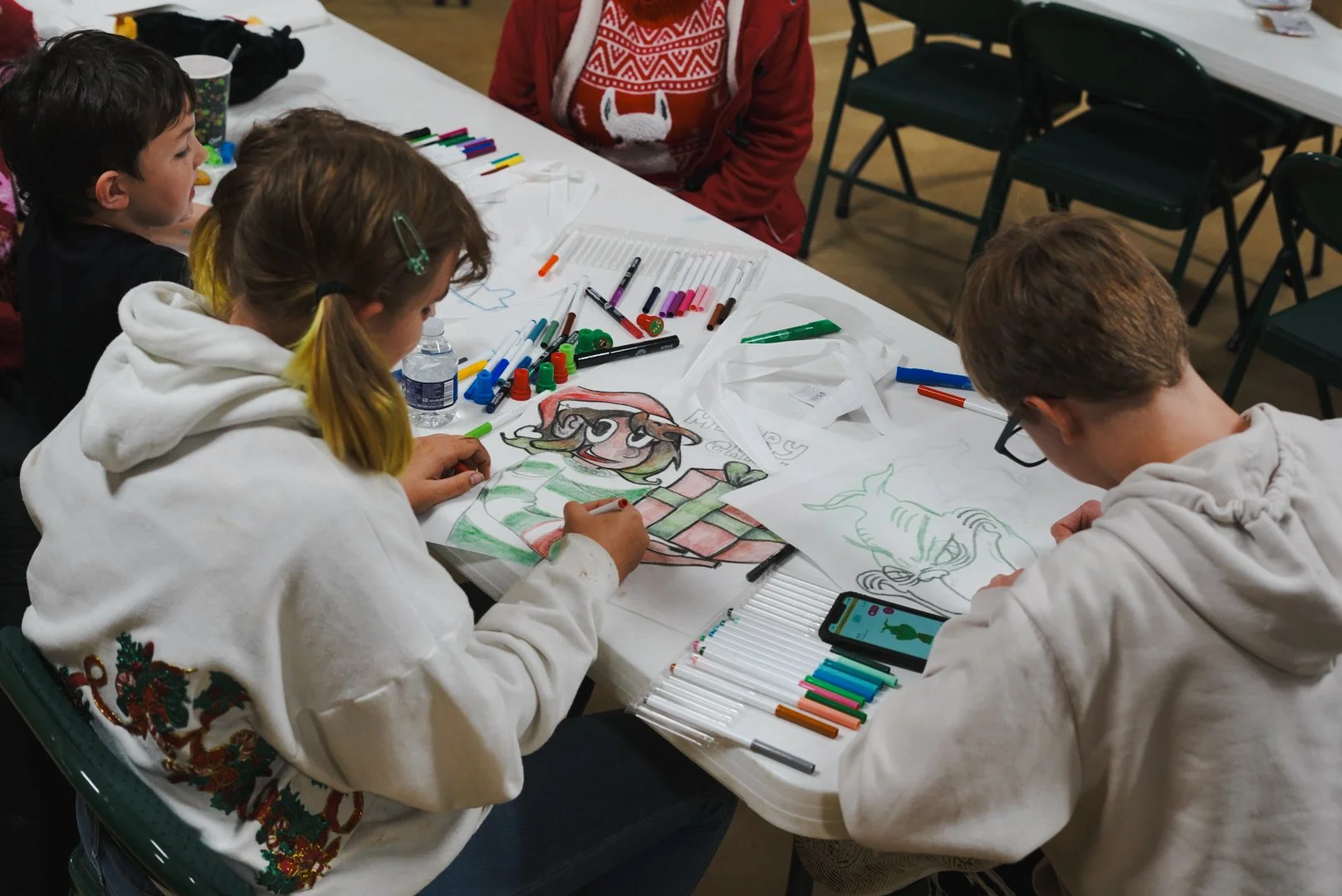 Children creating Christmas-themed holiday drawings at a table with markers and coloring supplies.