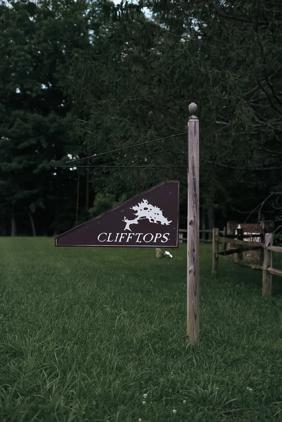 Wooden sign with a tree logo and the word 'CLIFFTOPS' hanging from a wooden post in a grassy area with trees in the background.