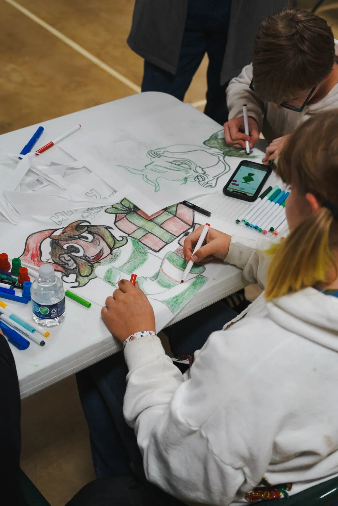 Children coloring holiday-themed characters on large sheets of paper, with markers, at a table.