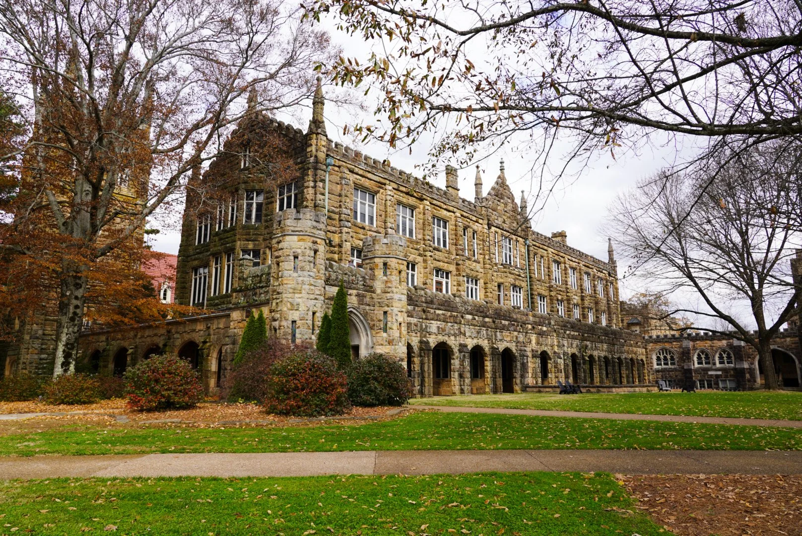 A historic stone building with multiple windows, surrounded by trees with autumn leaves and a grassy lawn with footpaths.
