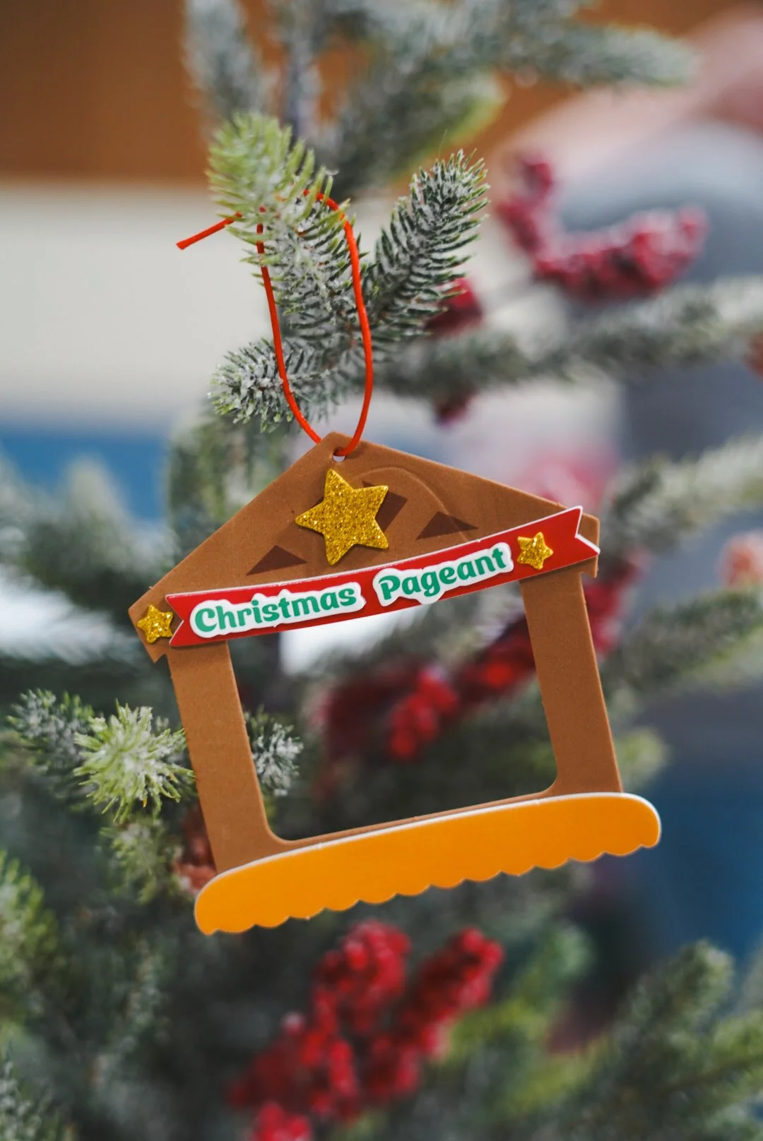 Christmas tree ornament in the shape of a picture frame with a sign that reads 'Christmas Pageant', hanging from a Christmas tree branch with snow-dusted green needles and red berries.