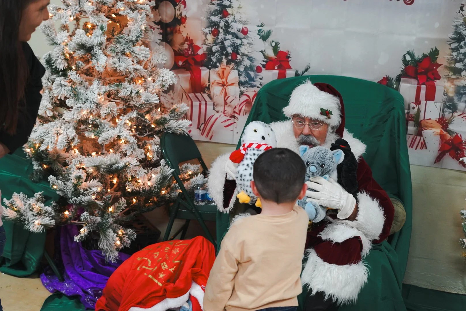 Santa Claus sitting on a green chair, holding stuffed animals, interacting with a young boy at a Christmas event with decorated trees and presents in the background.