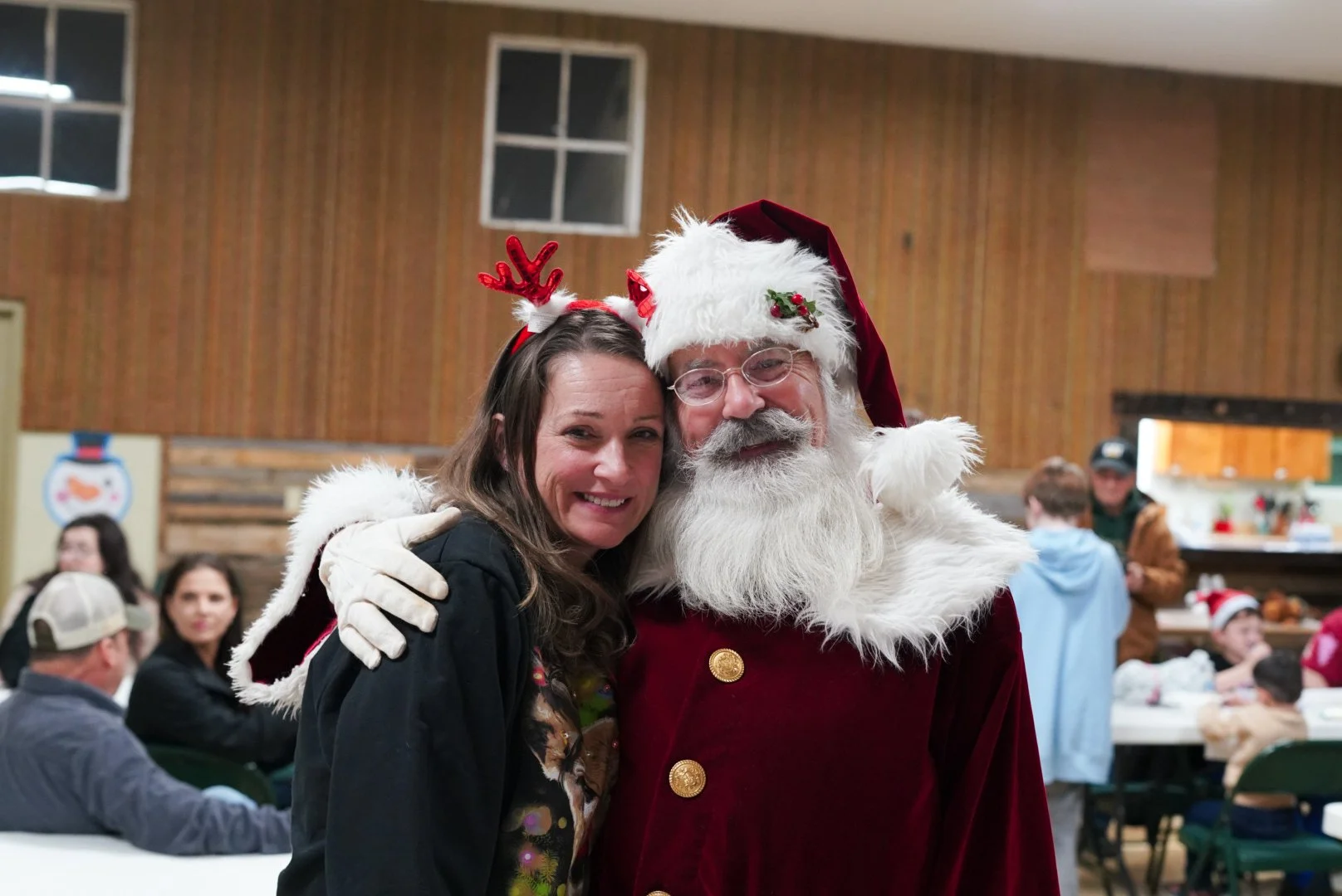 A woman and a man dressed as Santa Claus posing for a photo at a Christmas gathering, both smiling and embracing each other.