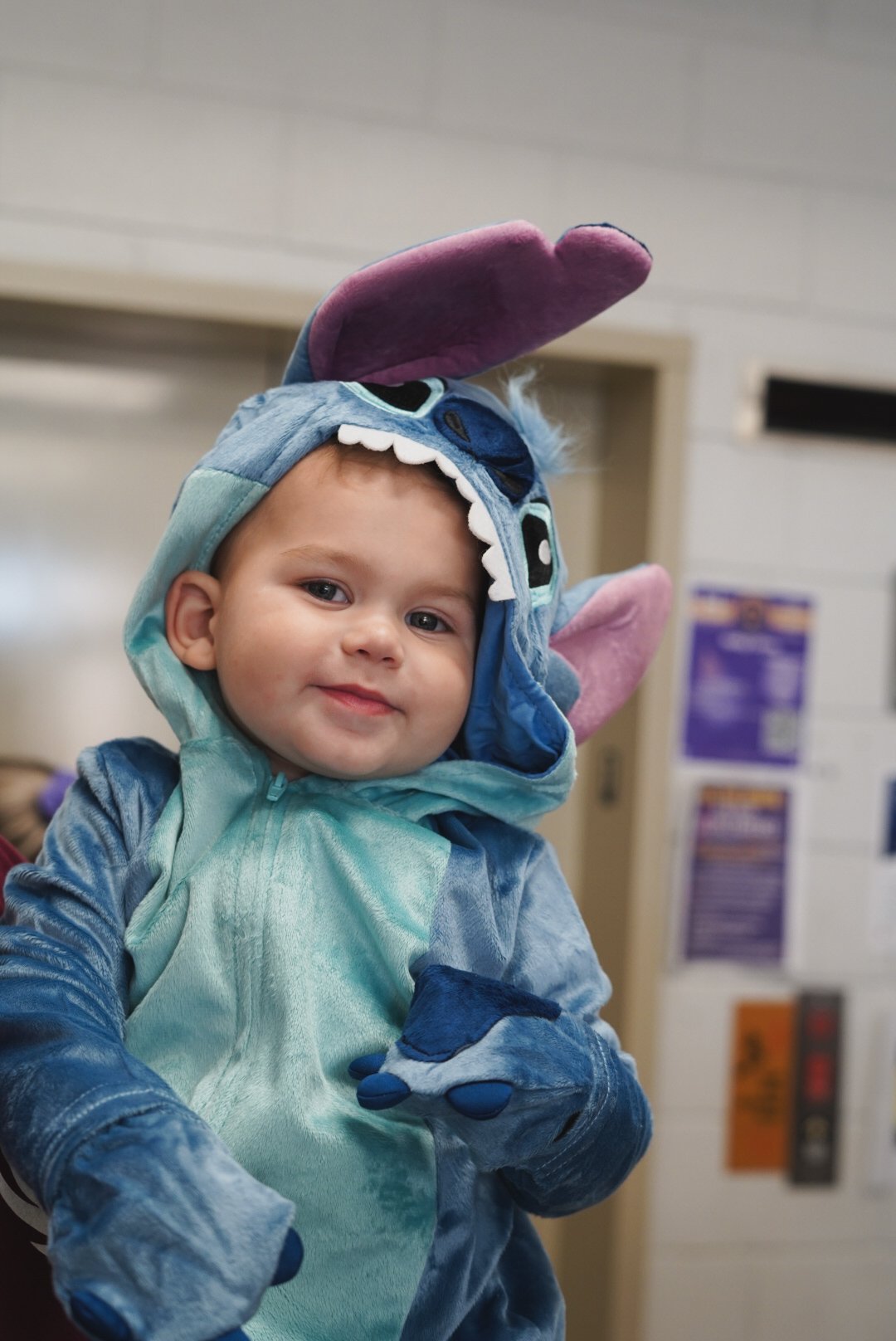 A young child dressed in a Stitch from Lilo & Stitch costume, complete with a hood featuring Stitch's face and large ears, smiling in an indoor setting.