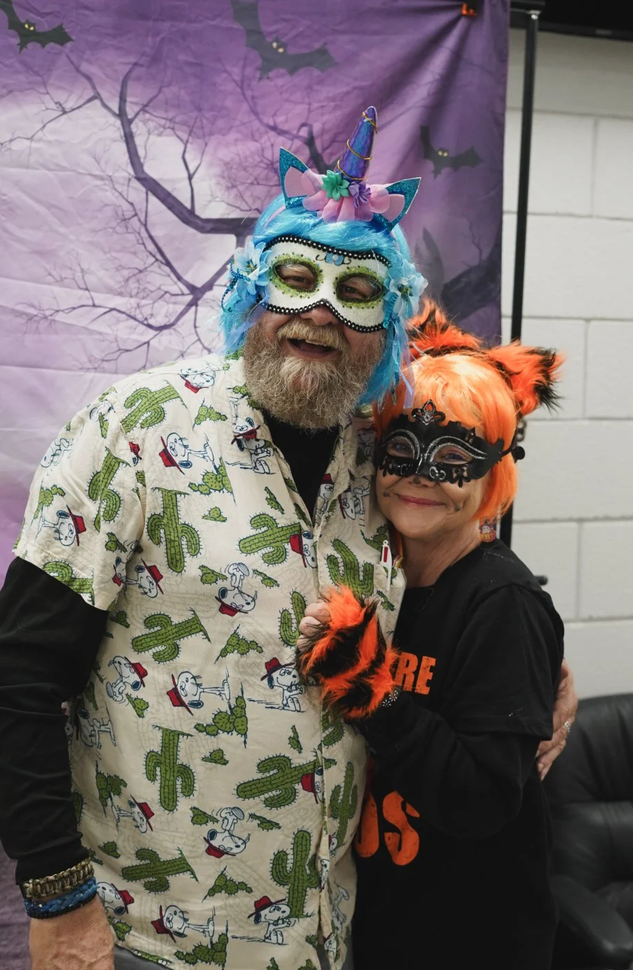 Two people wearing colorful costumes and masks, one with a blue wig and unicorn horn, the other with orange hair and fox ears, posing together in front of a Halloween-themed backdrop with bats and branches.