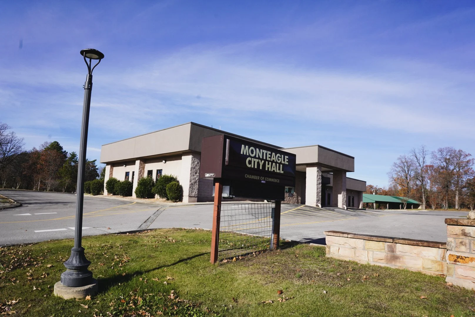 Exterior view of Monteagle City Hall with a black signboard reading 'Monteagle City Hall Chamber of Commerce', surrounded by bushes and an empty parking lot, under a clear blue sky.