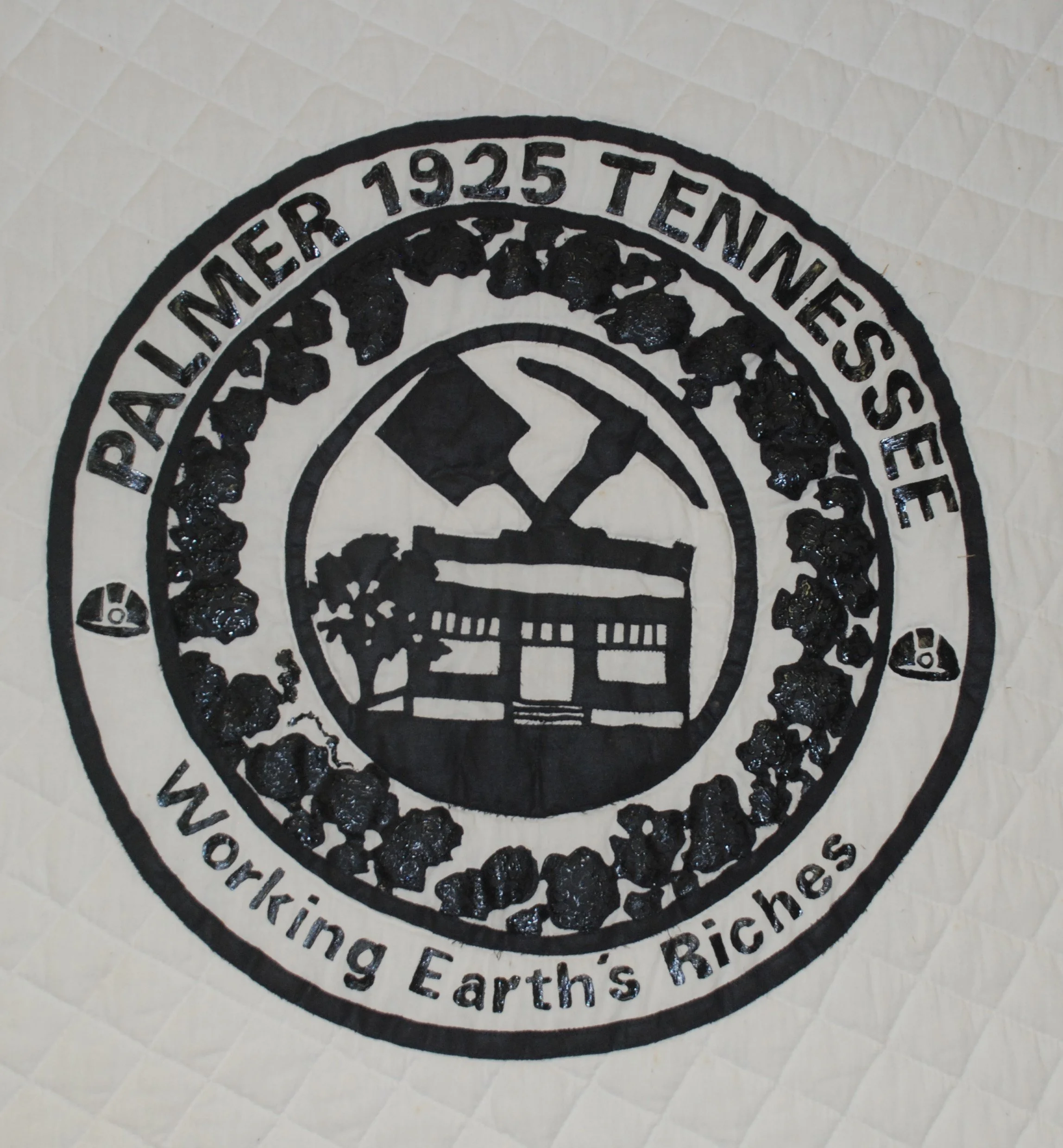 A circular logo with the text 'PALMER 1925 TENNESSEE' and 'Working Earth’s Riche' around the edge. The center features a black silhouette of a house with a tree and a mountain in the background, along with an axe and a hammer crossing each other.