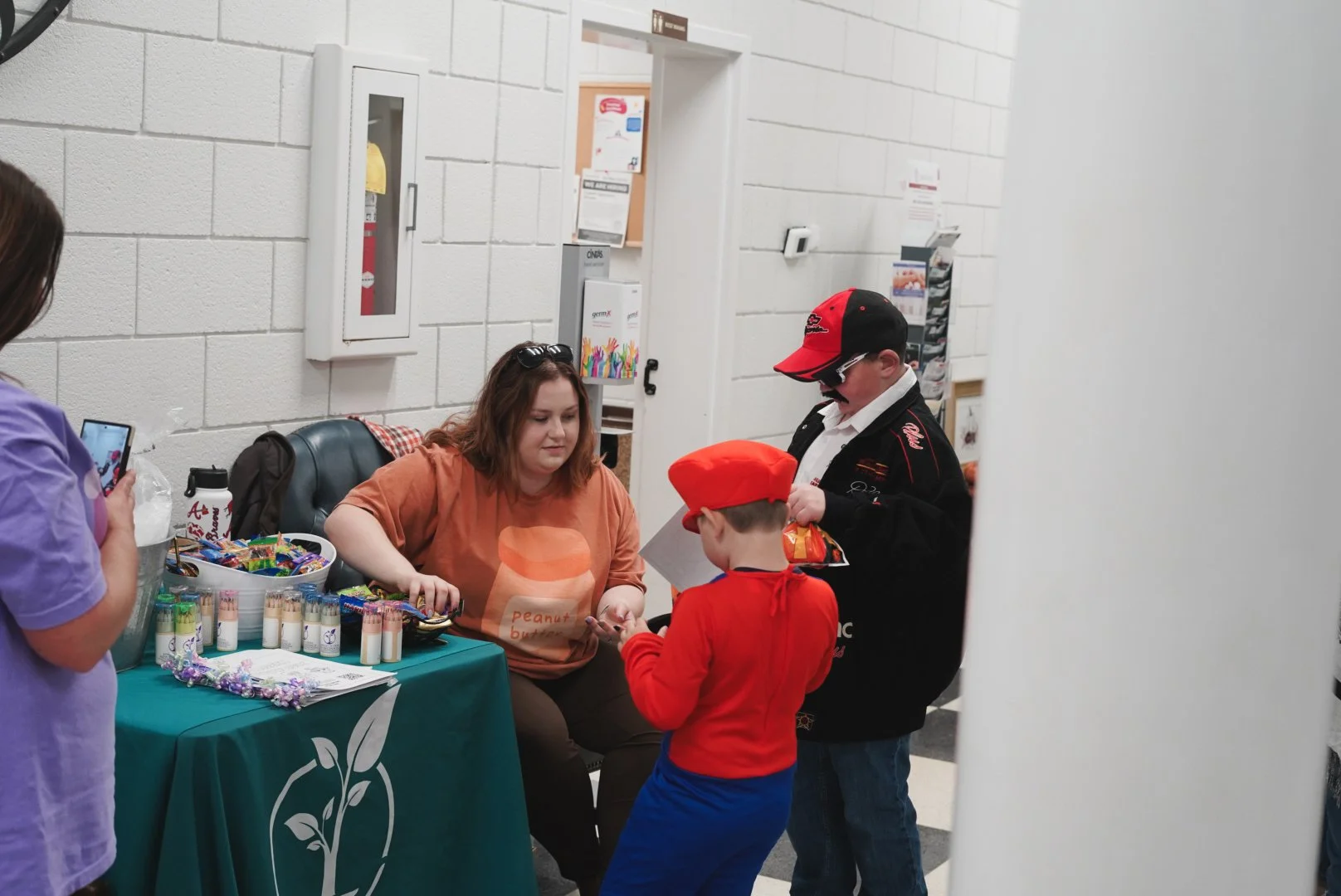 Woman sitting at table with candy and informational materials, children dressed as race car drivers talking to her, woman taking photo, parents and children in background.
