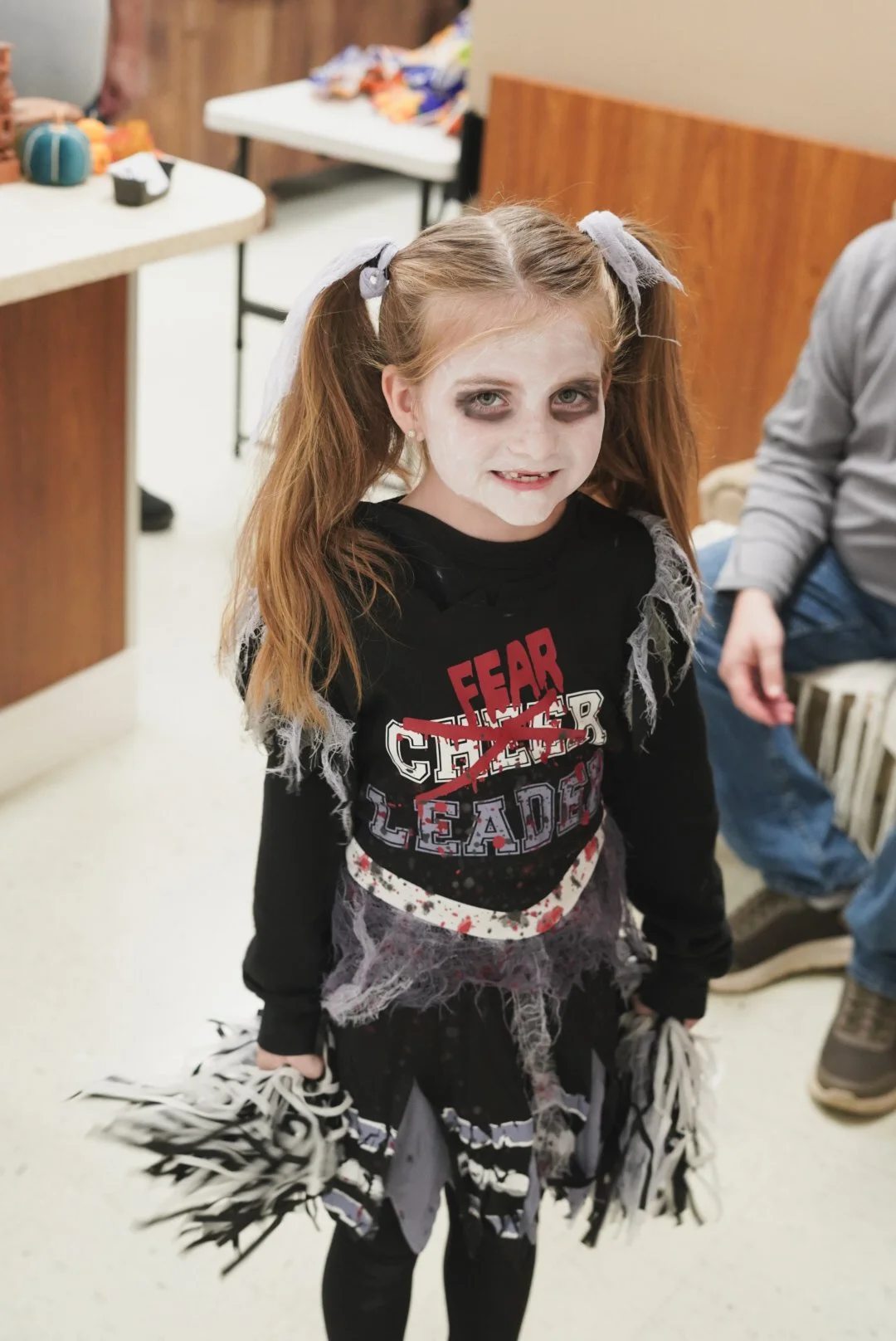 Girl dressed in a zombie costume holding pom-poms, with white face makeup and dark eye makeup, standing indoors near tables with snacks.