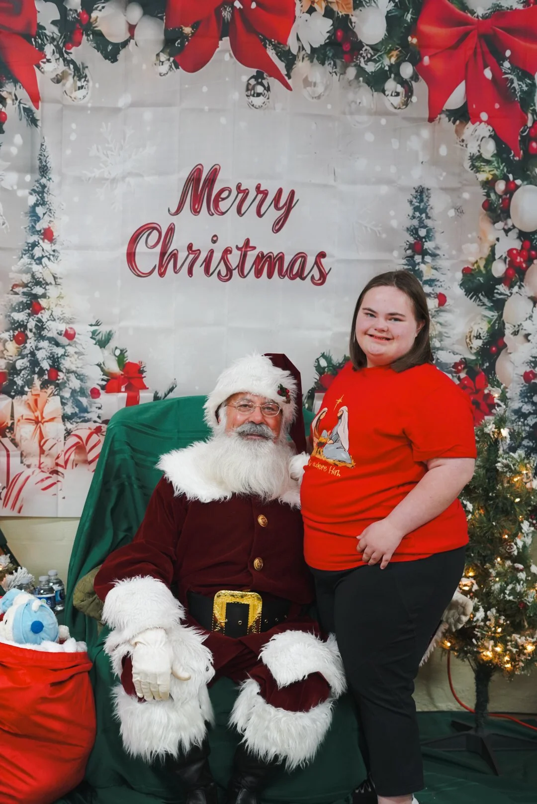 A young woman in red shirt standing next to Santa Claus in a festive Christmas setting with a decorated tree and "Merry Christmas" sign.