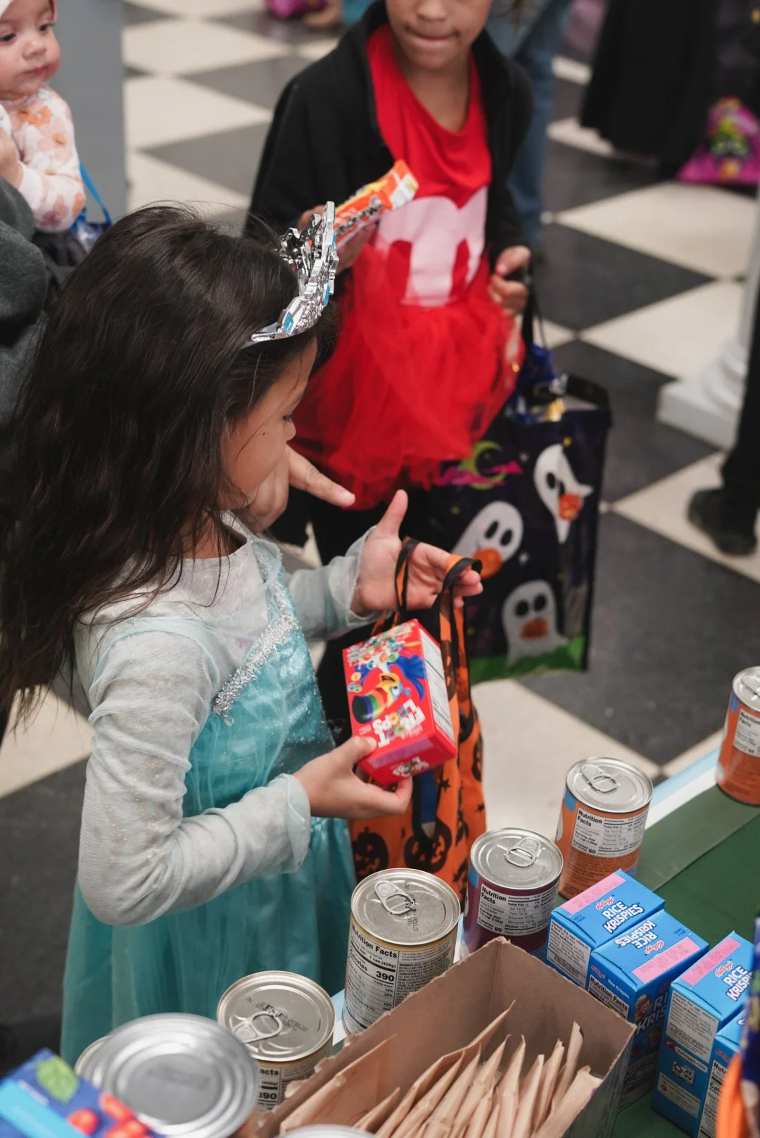 Children celebrating Halloween shopping, with a girl in a princess costume and others holding candy and paper bags, picking items at a grocery store.
