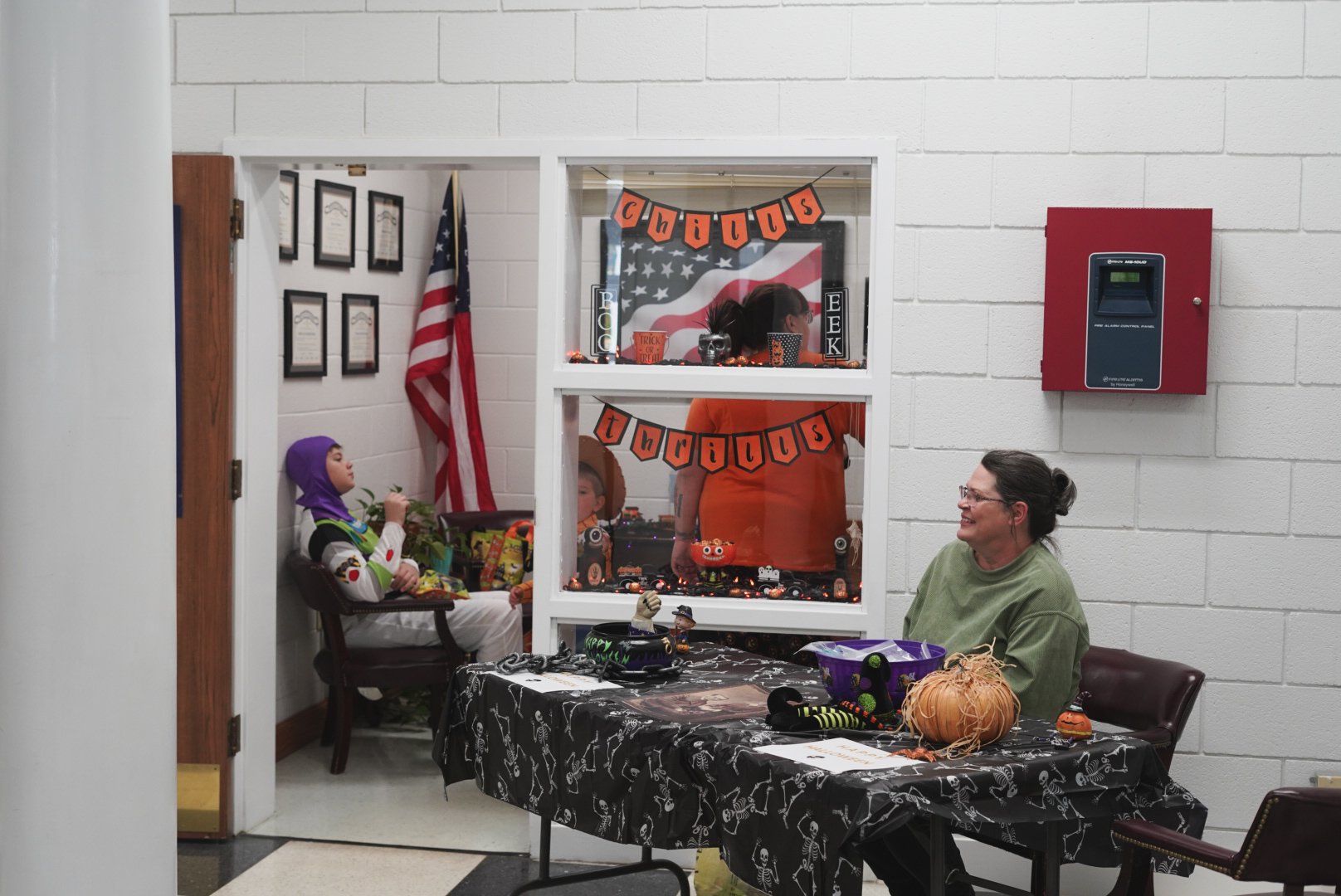 A woman sitting at a Halloween-themed table with pumpkins and decorations, smiling at a girl dressed as Buzz Lightyear sitting on a chair. Halloween decorations include a garland with the words 'chills & thrills,' Halloween lights, and table items. B