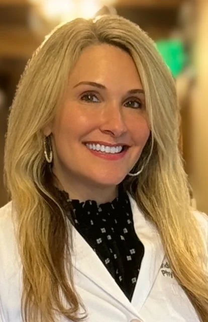 A smiling woman with long blonde hair, wearing a white lab coat and silver hoop earrings, indoors with blurred background.