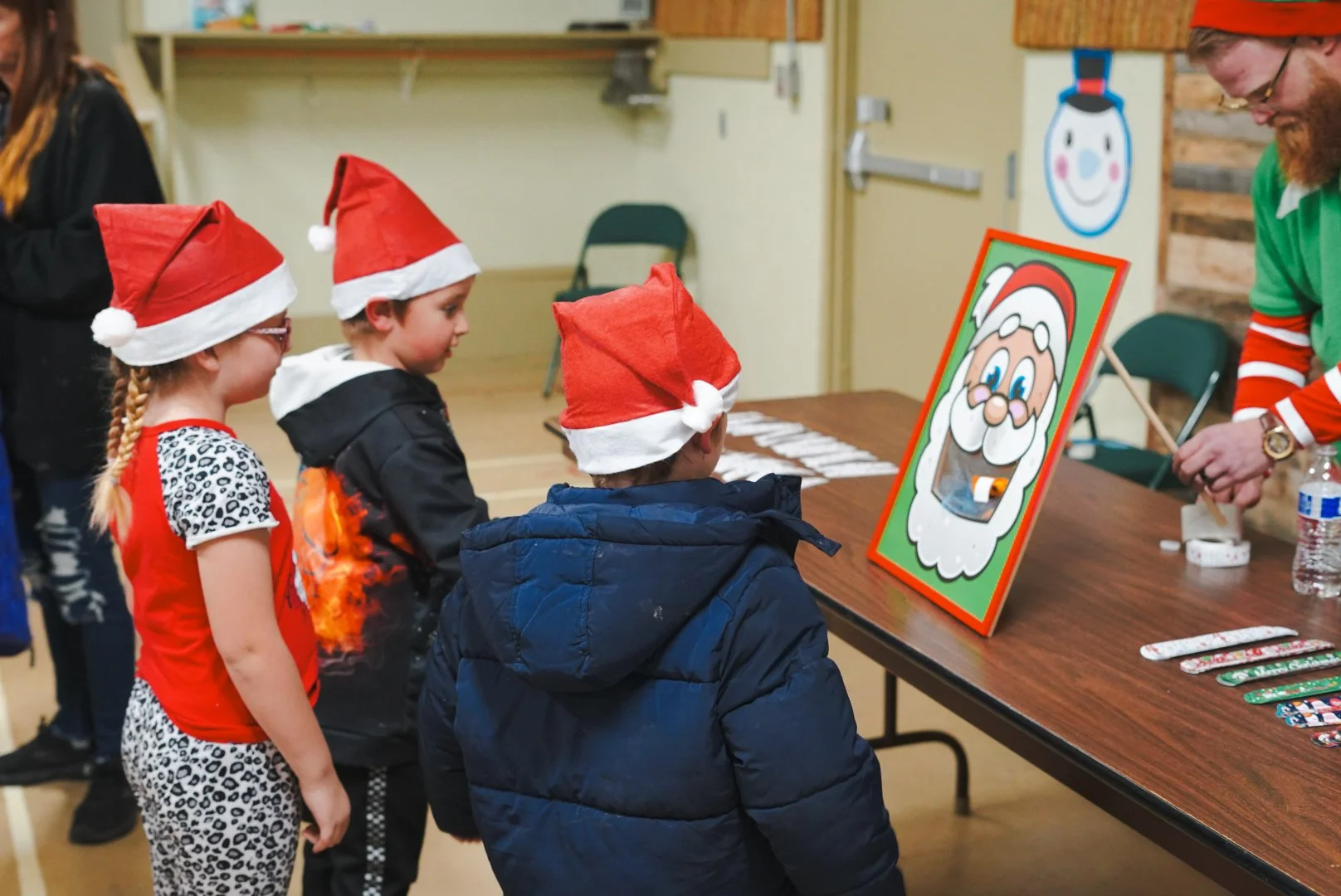 Three children wearing Santa hats standing in line at a table, waiting to meet a man dressed as Santa Claus, who is preparing to give out presents and wears a green holiday-themed shirt.