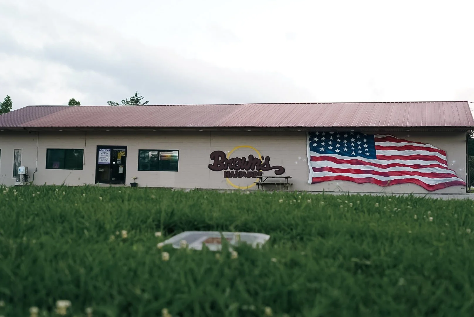 A beige building with a red metal roof, featuring a large American flag mural on the right side. The center displays a sign reading 'Brown's Hardware.' There are three windows and an entrance door, with some signs visible on the door. The foreground 