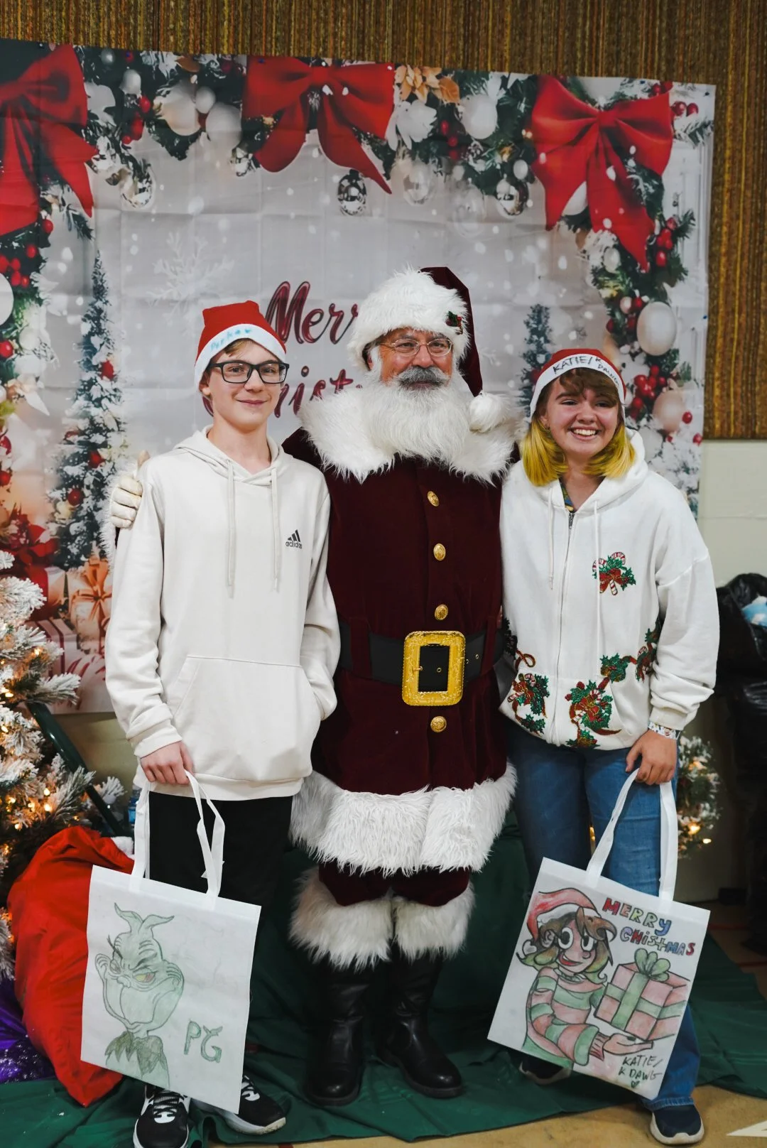 Two children, a boy with glasses in a white hoodie and a girl with yellow hair in a white jacket, stand next to a man dressed as Santa Claus in a festive setting with Christmas decorations and a backdrop that says "Merry Christmas." The boy holds a b