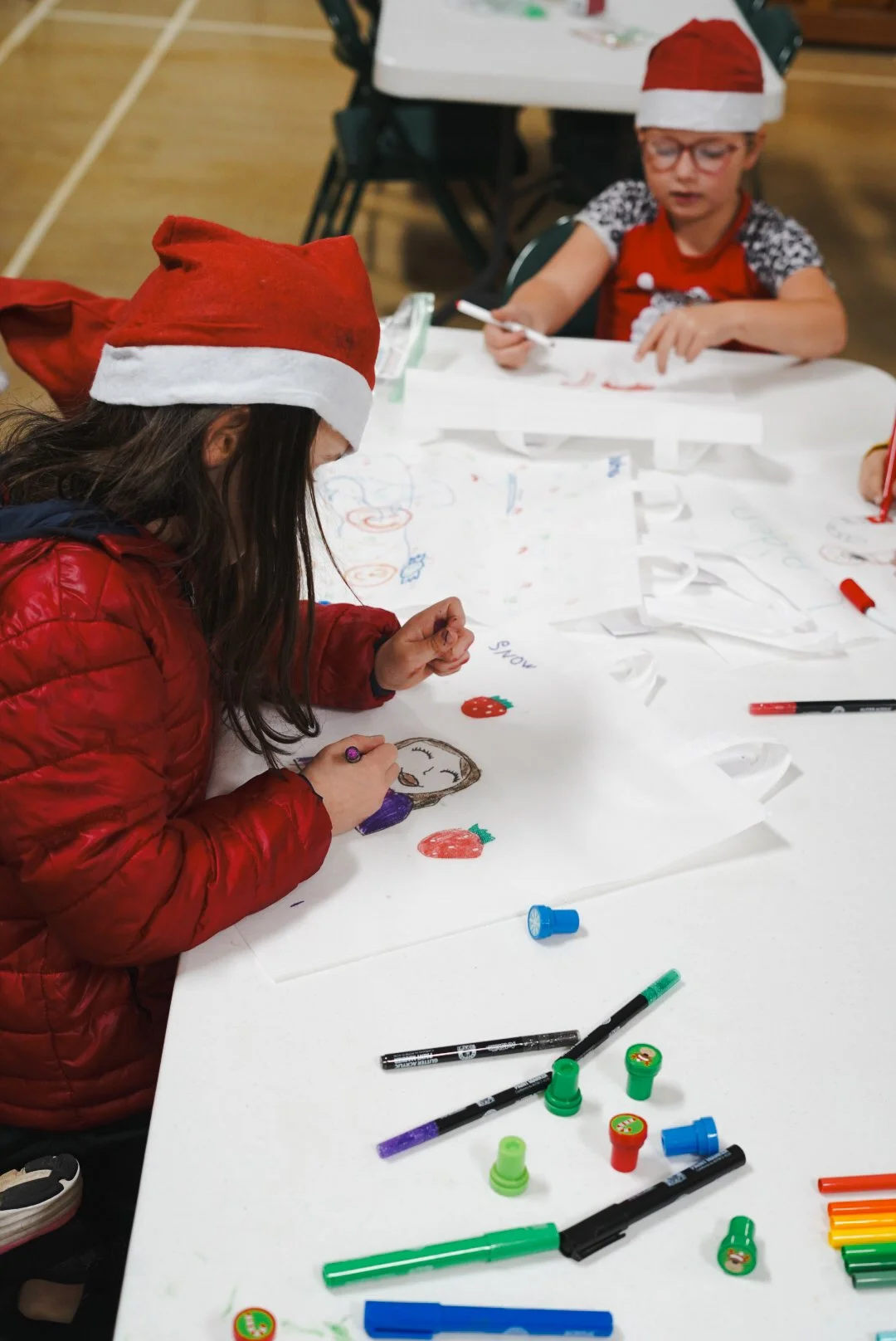 Children wearing Santa hats creating Christmas drawings at a table with markers and paper.