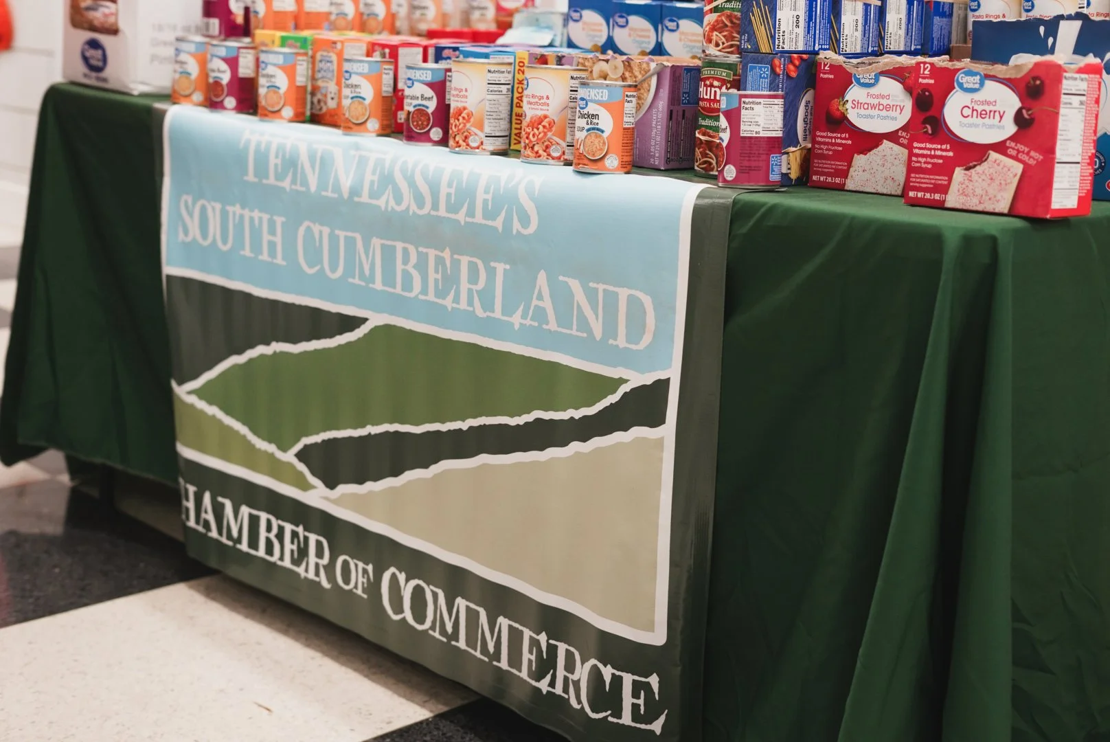 A table covered with a green tablecloth and a banner that reads 'Tennessee's South Cumberland Chamber of Commerce' displays various canned food products, including fruit and meat canned goods in a store or event setting.
