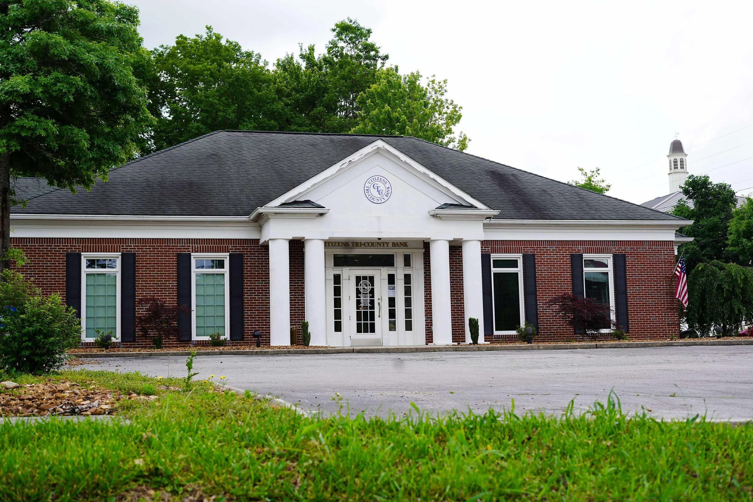 A brick bank building with white columns, black shutters on windows, and an American flag, surrounded by green trees and grass.