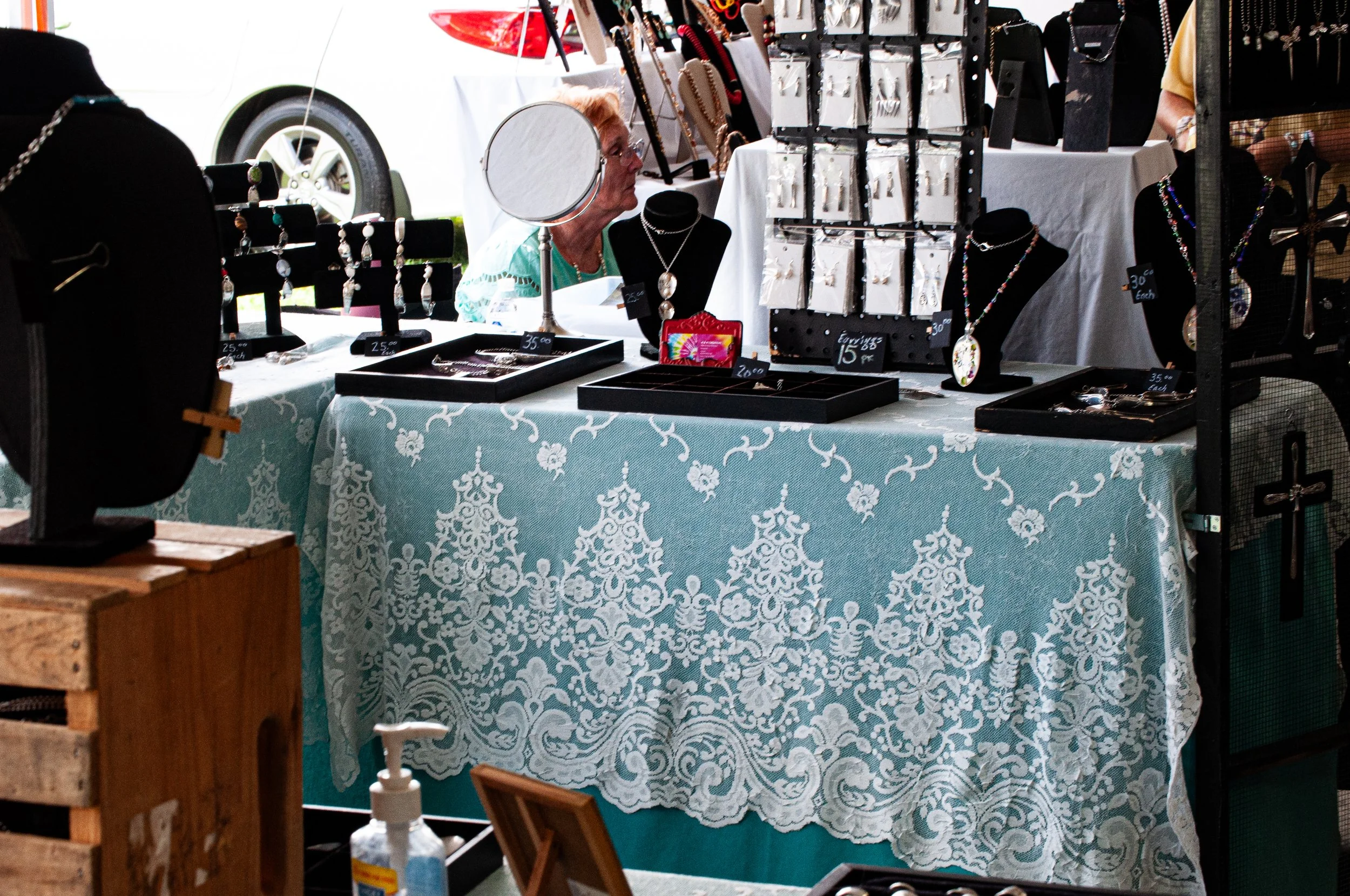A woman sitting behind a display table at an outdoor market. The table is draped with a blue and white patterned tablecloth and features jewelry including necklaces and earrings. Jewelry is displayed on black stands and in trays with price tags. To t