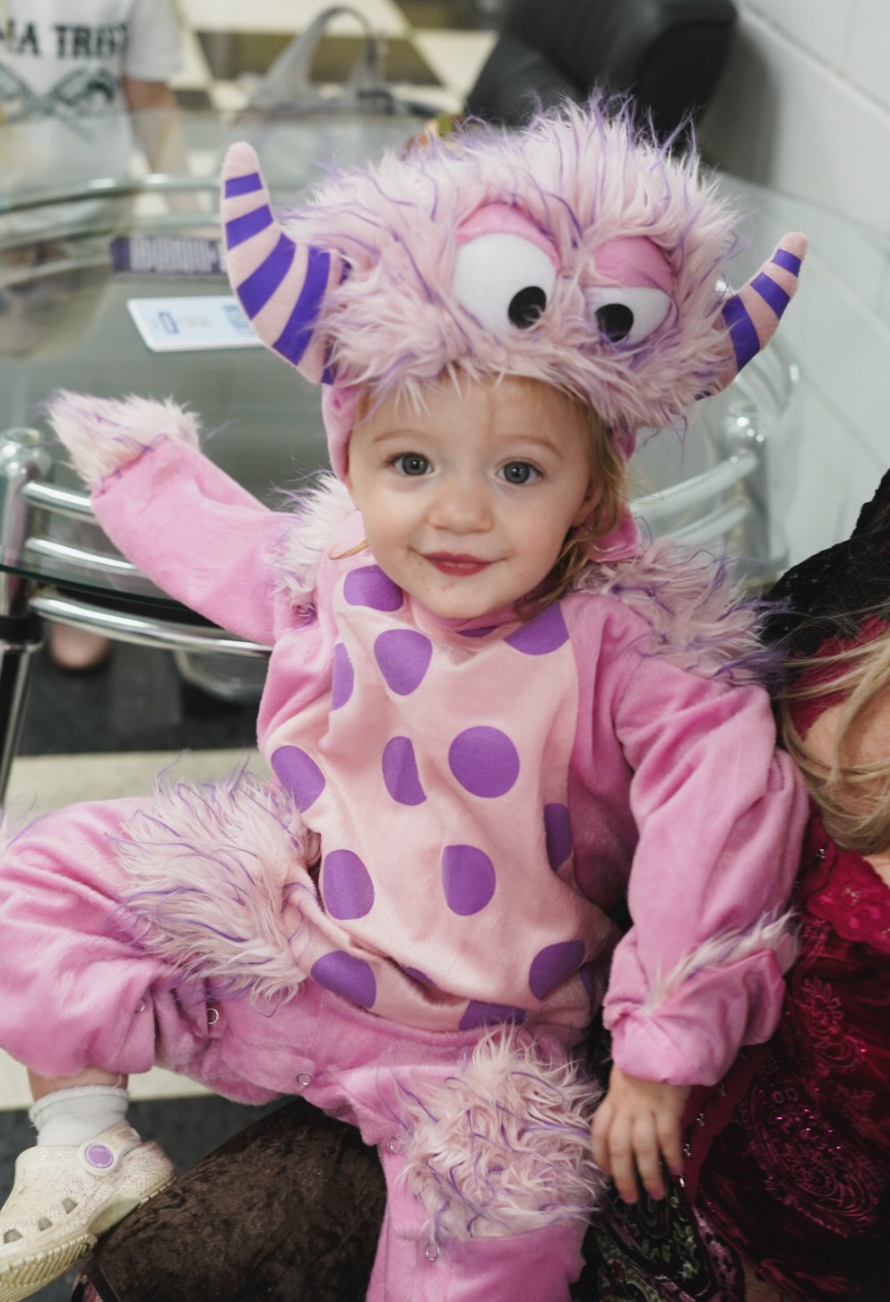 A young girl dressed in a pink monster costume with purple polka dots, furry details, and a headpiece resembling a friendly monster with big eyes and striped horns.