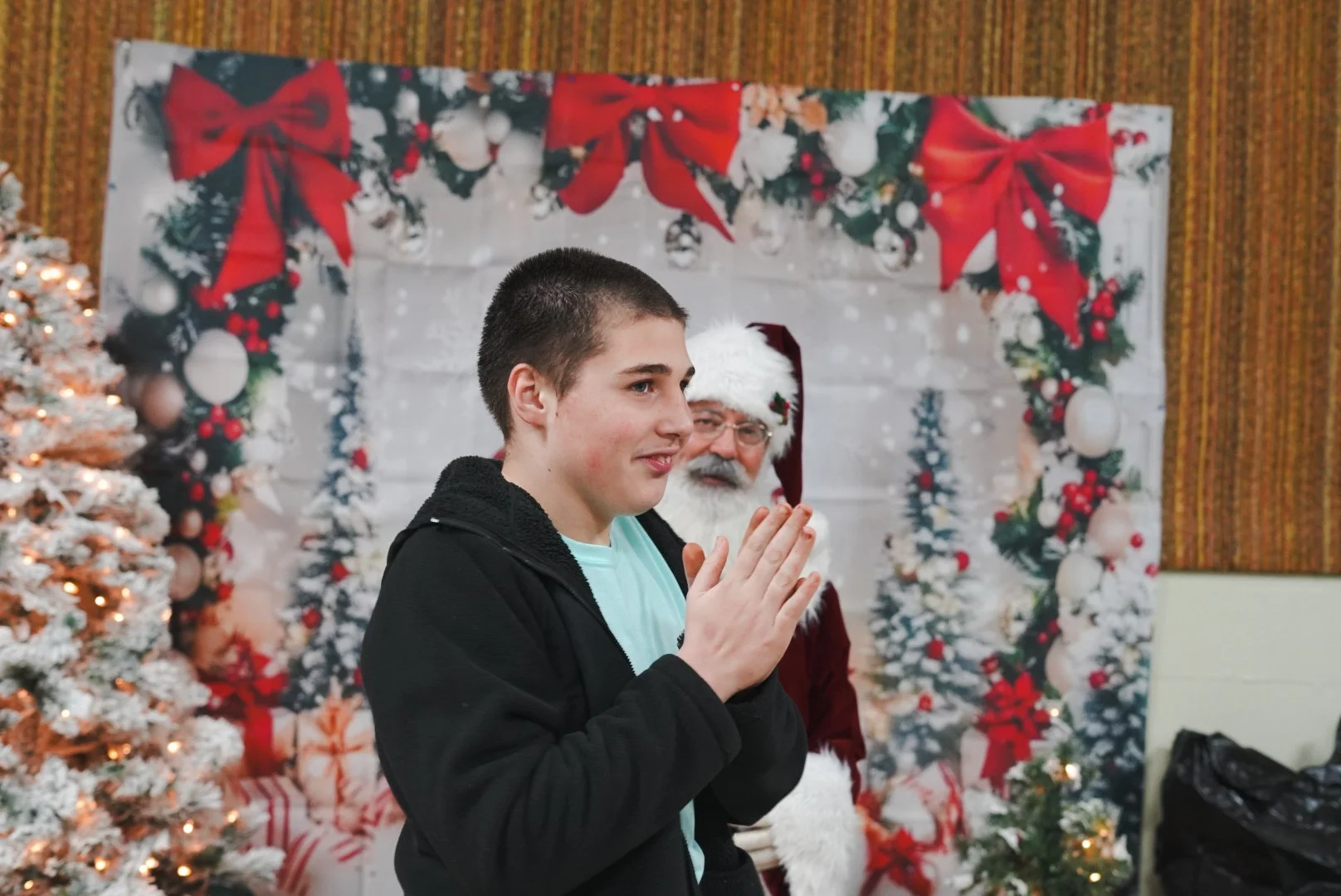 A young man with short dark hair and a light blue shirt standing with his hands together in front of a Santa Claus in a festive holiday setting. Santa is wearing a red hat with white trim, glasses, and has a gray beard. Behind them is a decorated bac
