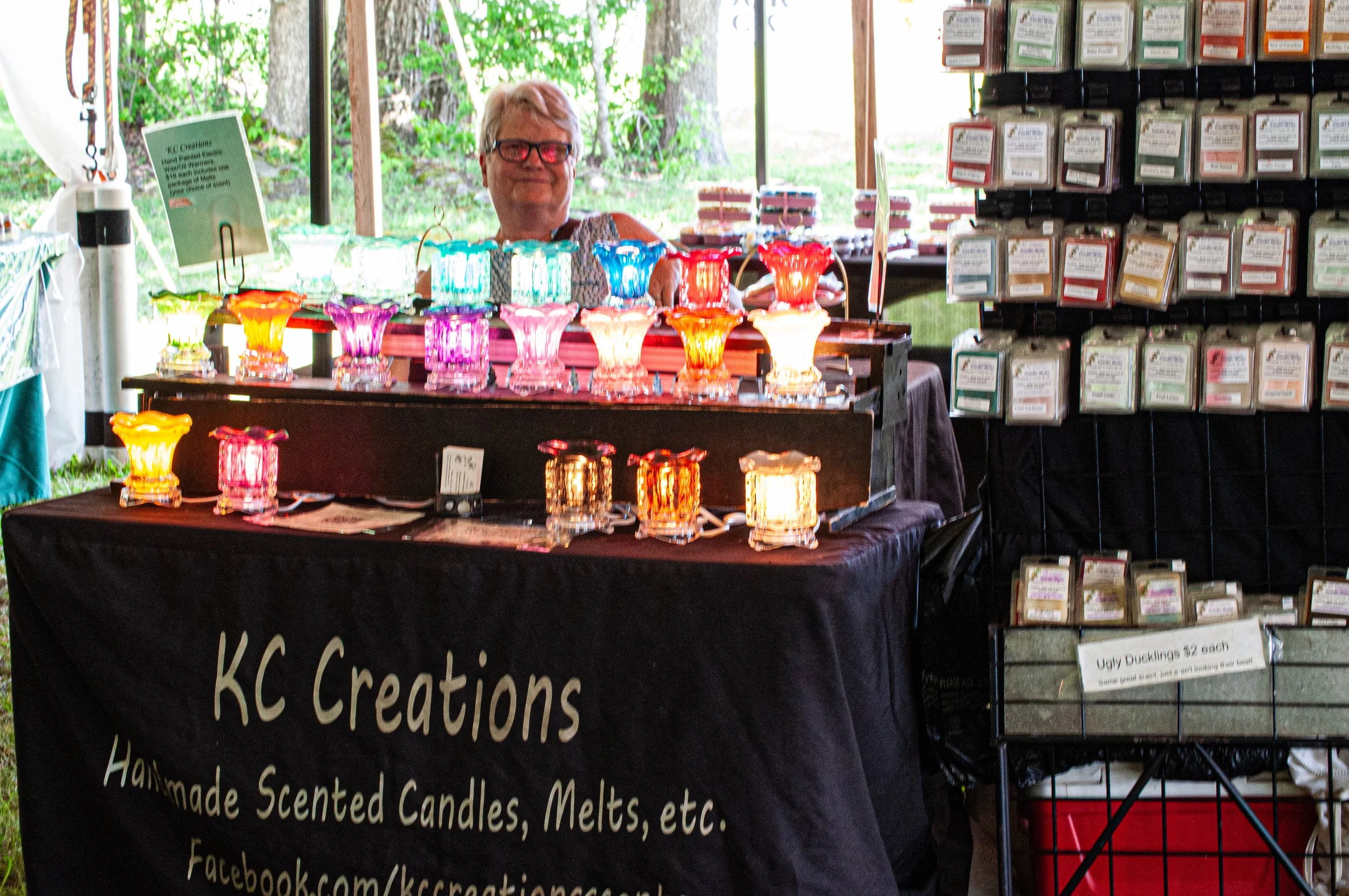 A smiling person with glasses behind a display of colorful handmade candles at an outdoor craft fair. The candles are in various bright colors and shaped like flowers. On the right, there are shelves with packaged scented candles and other items.
