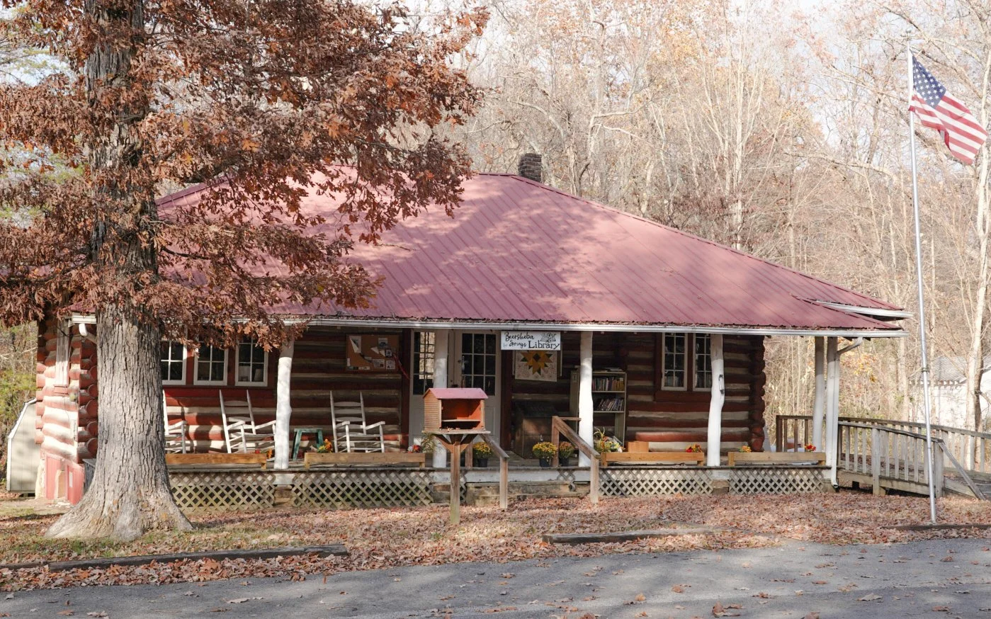 A log cabin-style building with a red metal roof, surrounded by trees with autumn leaves. The building has a porch with rocking chairs, an American flag on a flagpole, and a small sign that reads “Beersheba Springs Library”.