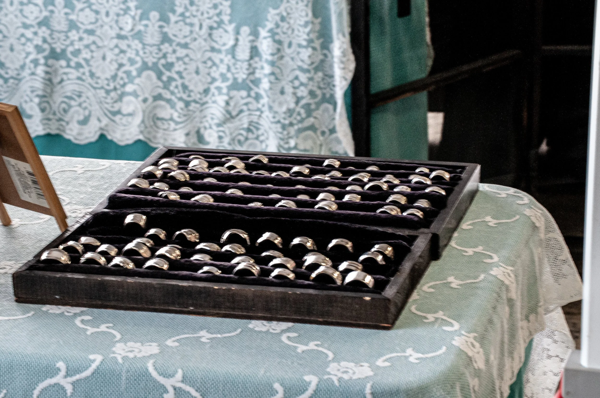 Box of silver rings on a black display tray set on a table with a lace tablecloth.