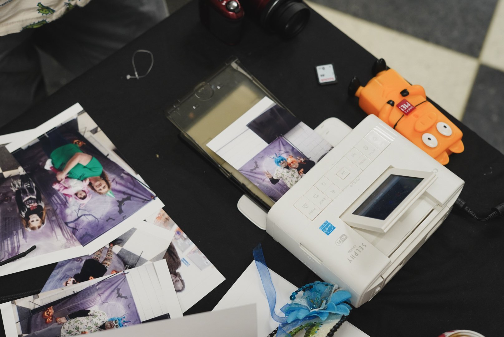 A black table holding a photo printer, printed photos, a camera, a small orange toy, and various electronic items.