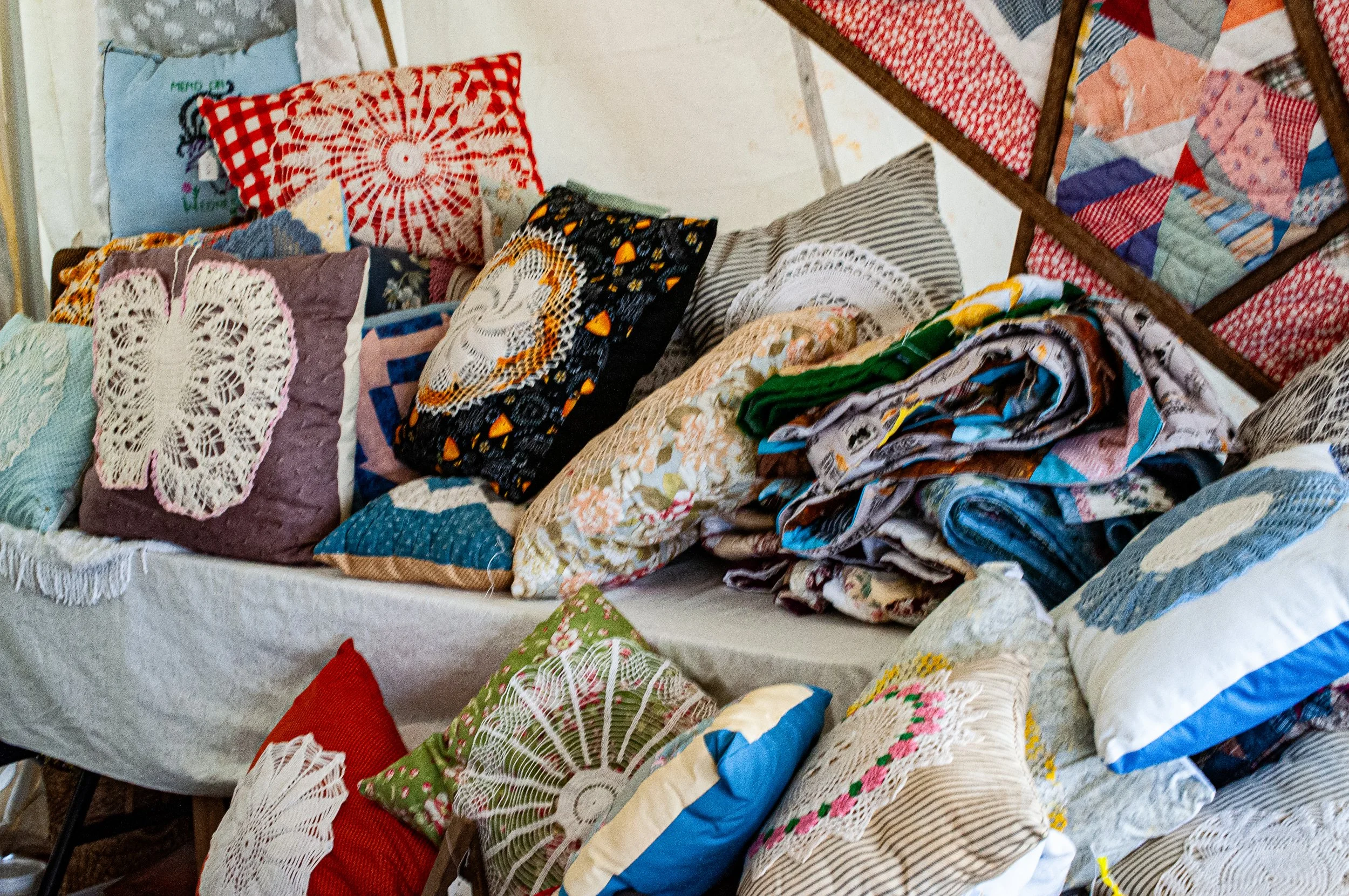 A collection of decorative pillows and fabric pieces with various patterns and crochet details, displayed on tables and chairs, creating a colorful and textured scene.