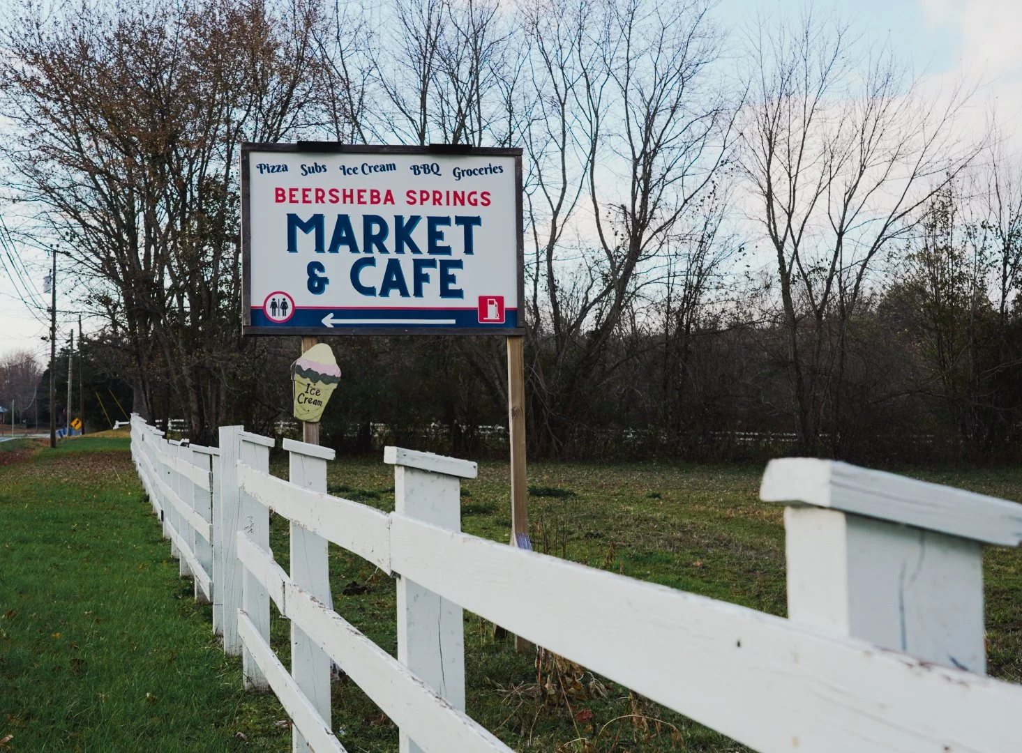 Sign for Beersheba Springs Market & Cafe with additional signs for pizza, subs, ice cream, BBQ, groceries, and ice cream nearby, along a white fence on a rural road.