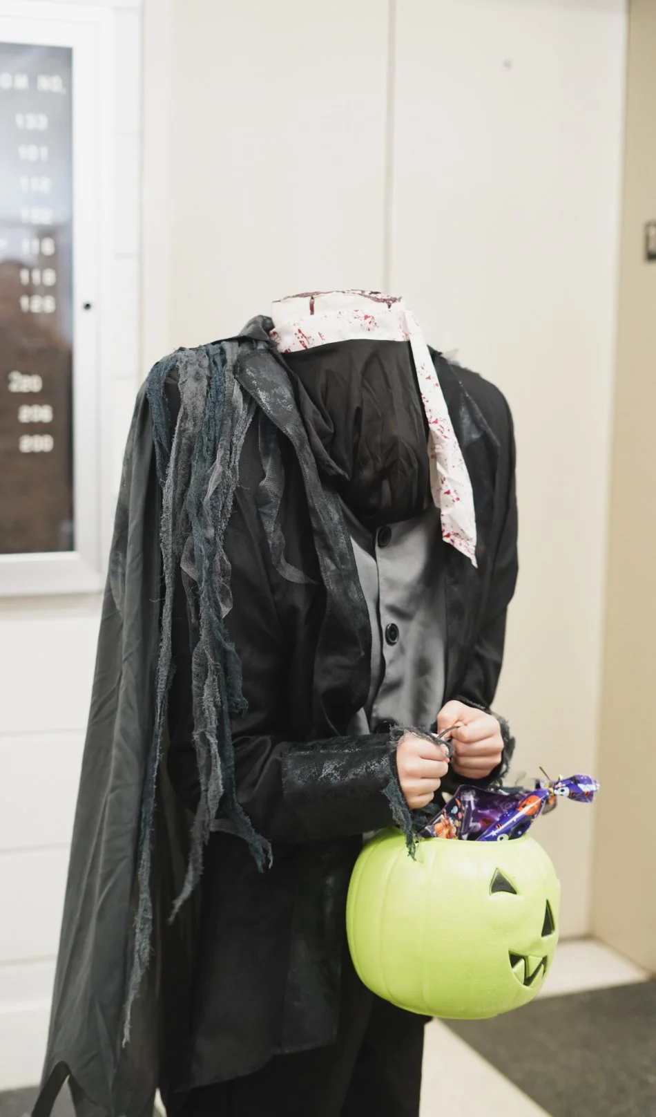 A person dressed in a Halloween costume with a tattered black cloak, a blood-splattered white cloth over the face, holding a green pumpkin-shaped bucket filled with candy, standing in front of an elevator at a building.