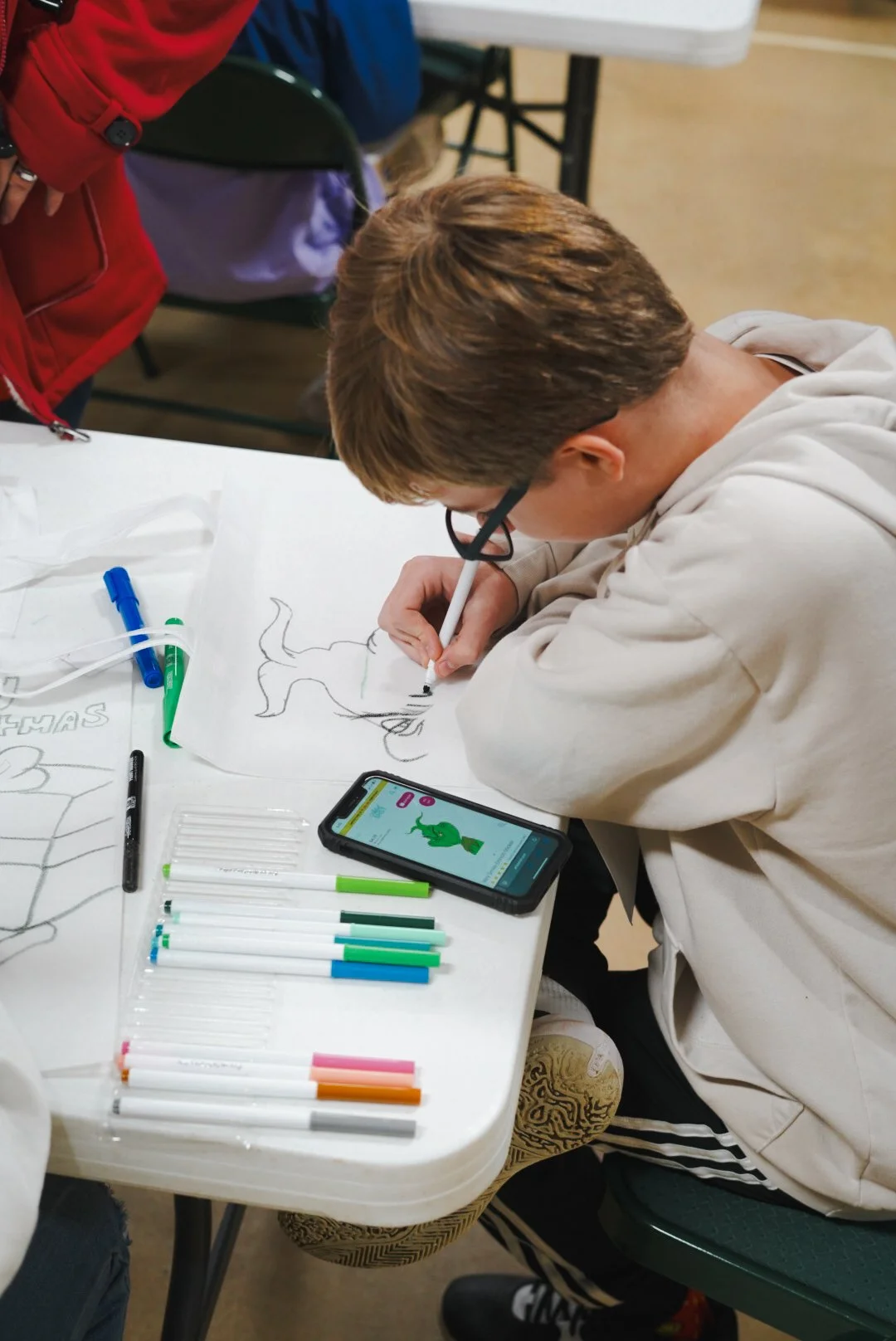 A young person drawing a dinosaur on paper with markers on a table, surrounded by pens and a smartphone displaying a dinosaur illustration.