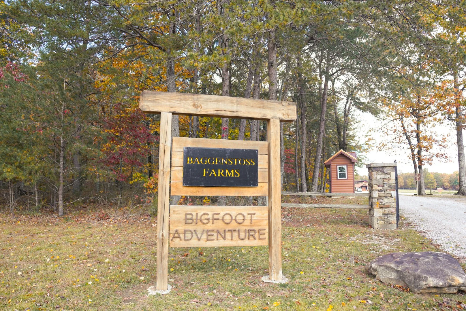 A wooden sign at the entrance of Baggerstoss Farms, advertising Bigfoot Adventure, with trees and a gravel road in the background.