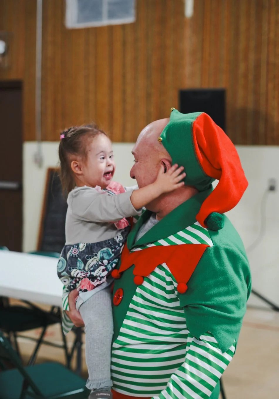 A young girl smiling and reaching out to touch the face of a man dressed as an elf, who is holding her. The elf costume is green with red accents, a large hat, and striped shirt, and the scene appears to be in a festive indoor setting.