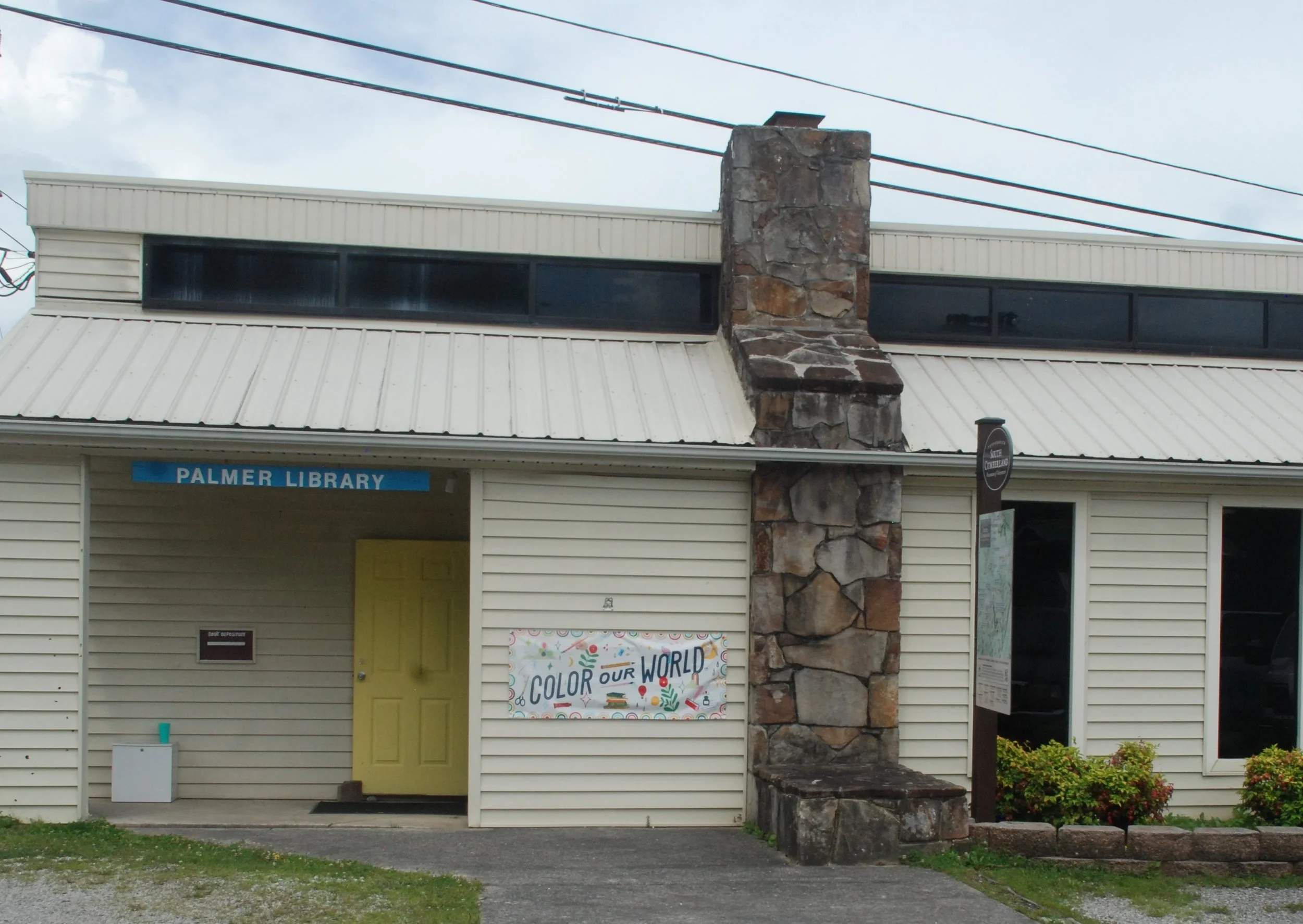 The exterior of Palmer Library building with a yellow door, a stone chimney, and a colorful sign that reads 'Color Our World.'