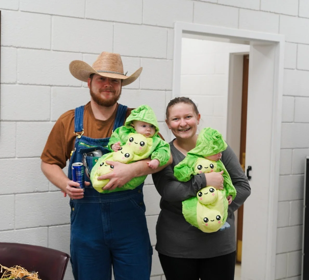 A man and woman holding two babies dressed in matching lime green avocado-themed costumes with smiling faces, standing in front of a white brick wall.