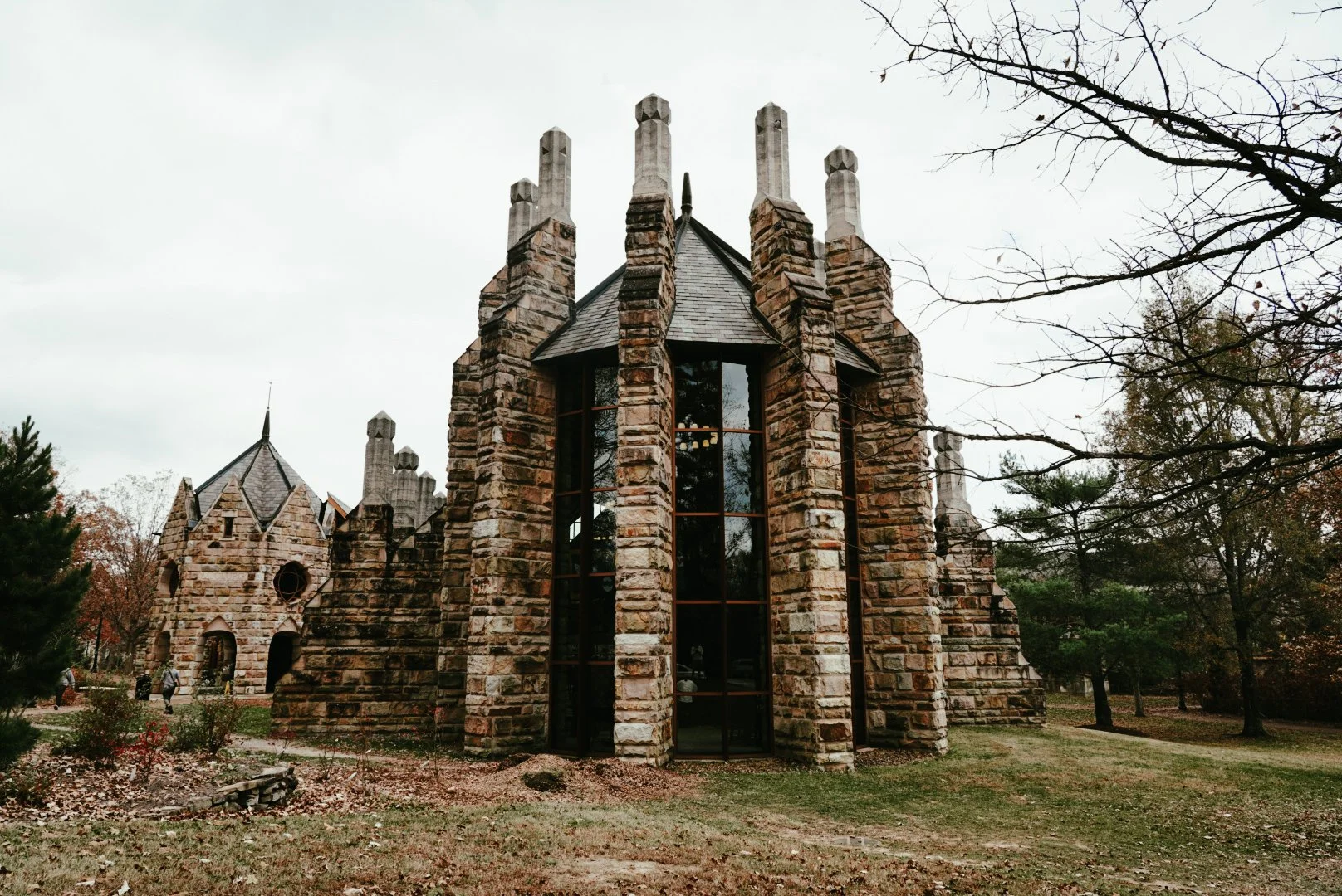 A stone castle-like building with tall, pointed towers and narrow windows, surrounded by trees and grass on an overcast day.