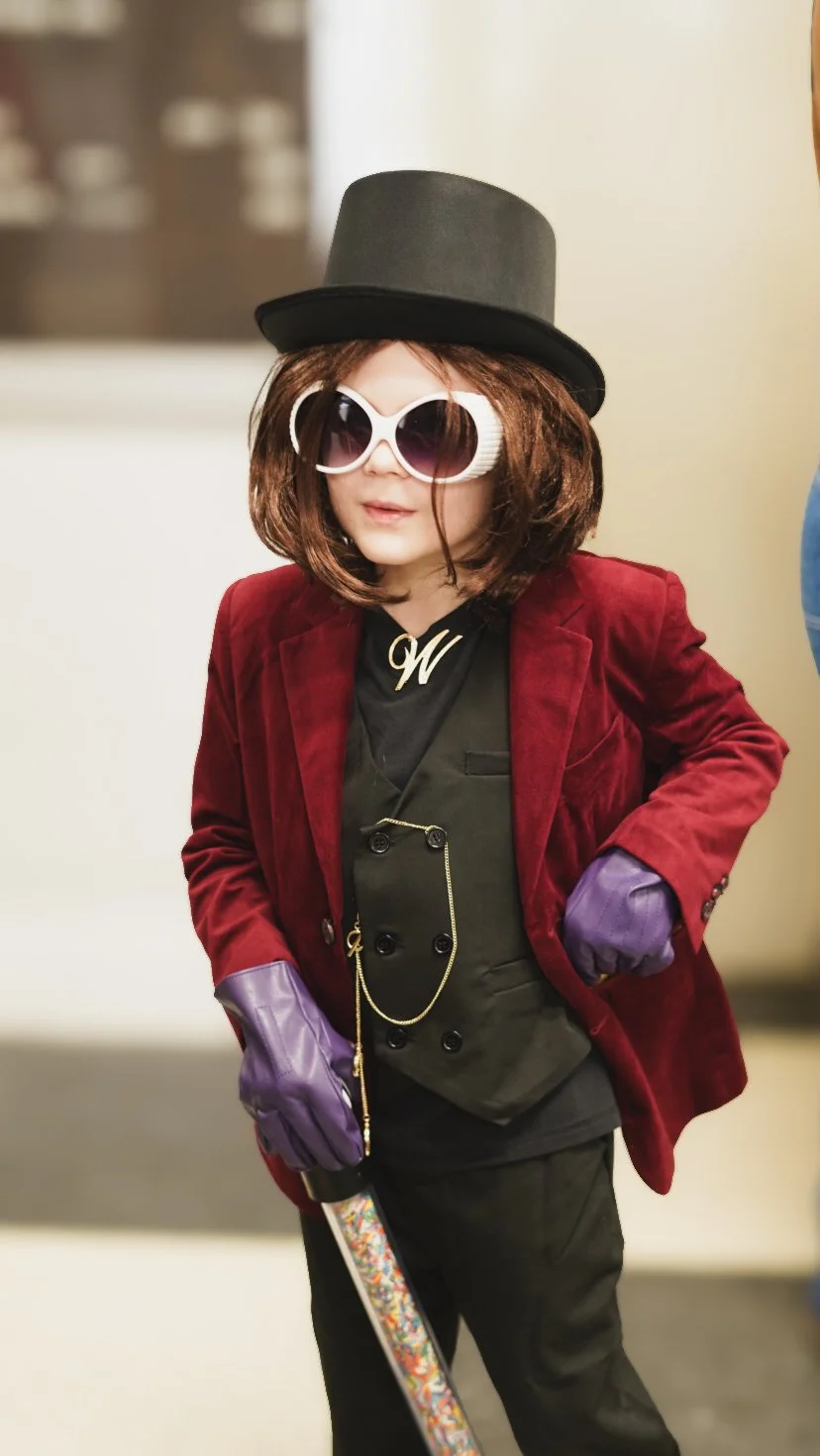 Person dressed in a flamboyant costume with a black top hat, large sunglasses, purple gloves, a red velvet blazer, a black vest, and black pants, holding a colorful cane.