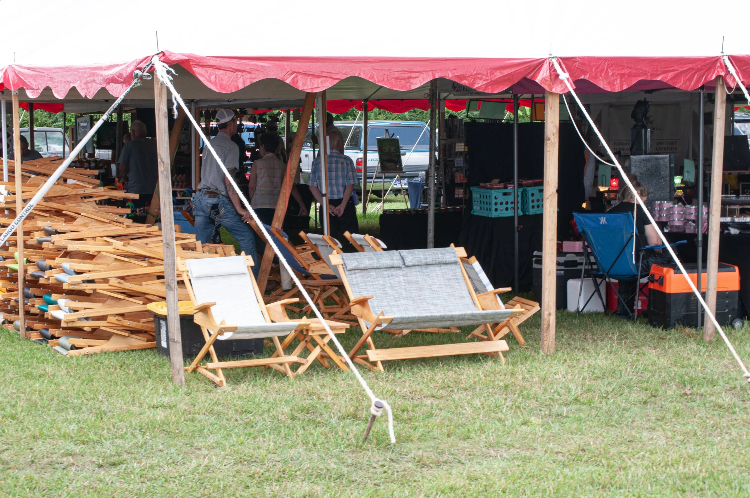 Outdoor market tent with people browsing and chairs and wooden items displayed in front.
