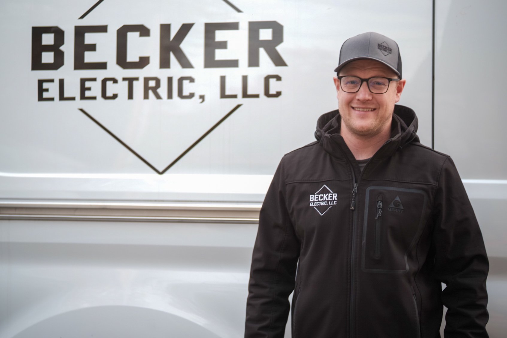 A smiling man in a black jacket and gray baseball cap standing in front of a Becker Electric LLC vehicle with logo