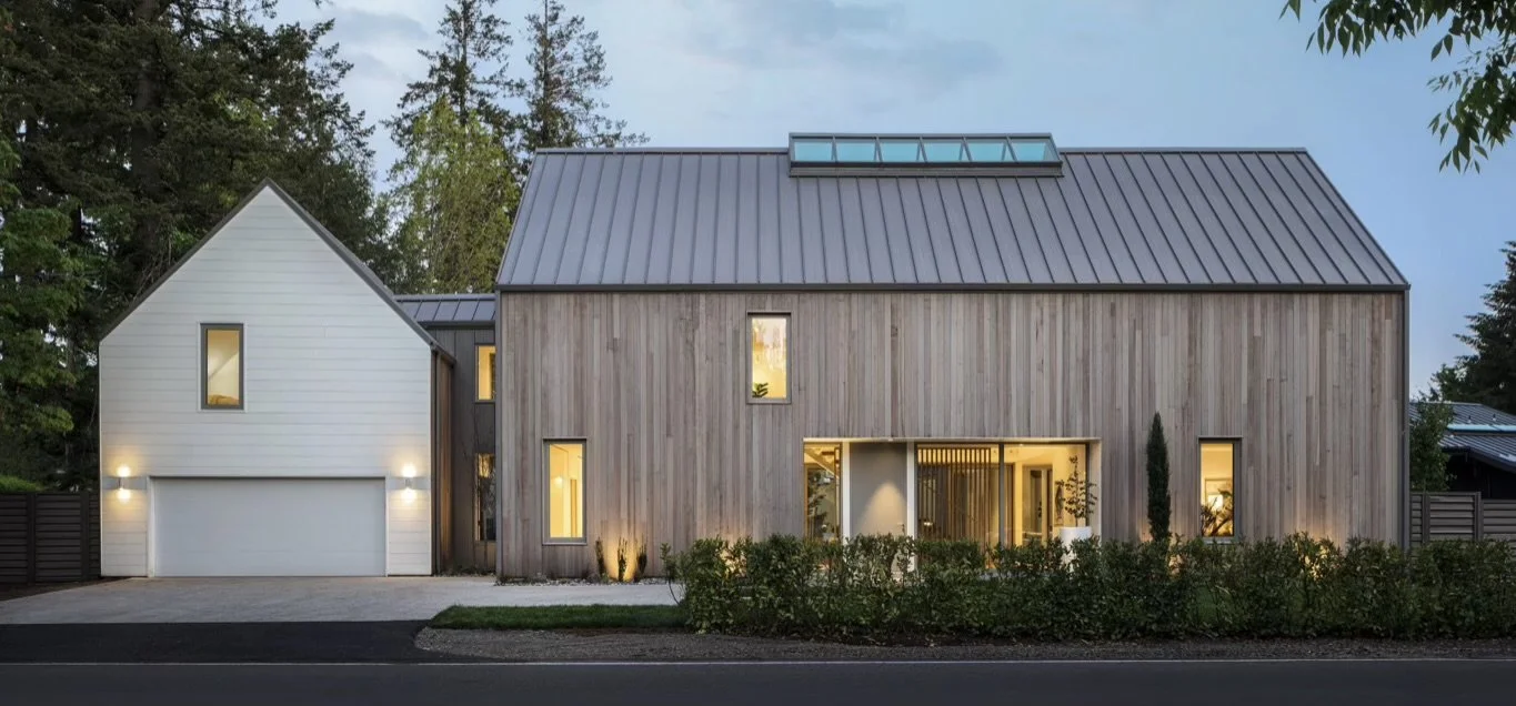 Modern house with a combination of white and wooden exterior, metal roof, and a garage, situated along a street with landscaping.