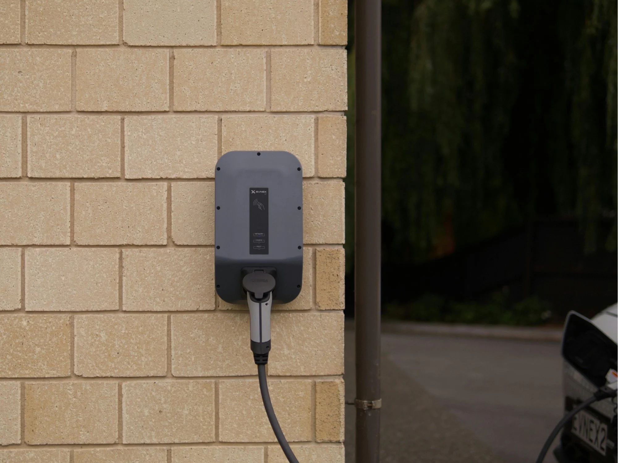 Electric vehicle charger mounted on a brick wall with a connected charging cable, situated outdoors near a parking area.